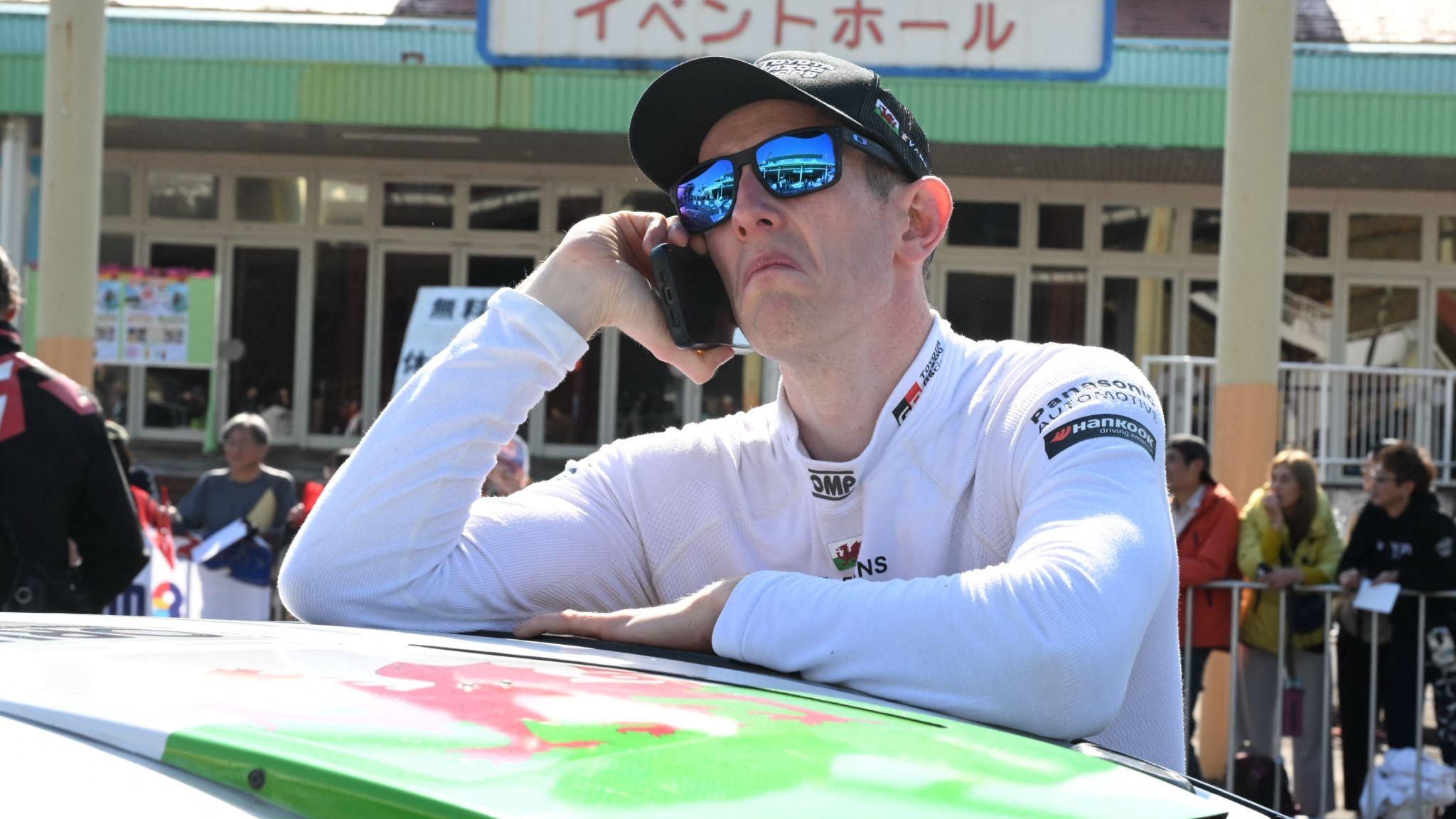 Elfyn Evans wearing a cap and sunglasses talks on the phone while leaning on the roof of a car painted with a Welsh flag.