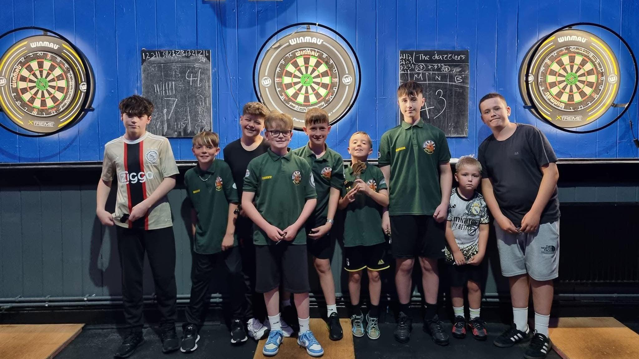 Members of Shooters Leominster Junior Darts Academy, wearing green club T-shifts, shorts and trainers, with darts boards behind them.