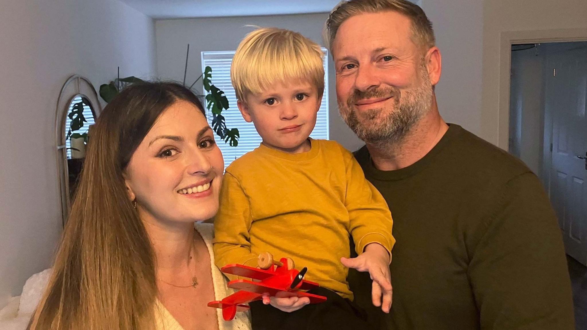 Monika, Henry and Leo smile at the camera in a living room. Monika, who has long brown hair, and Henry who has blonde short hair and a brown jumper are holding toddler Leo who is holding a red toy plane and has a yellow jumper on