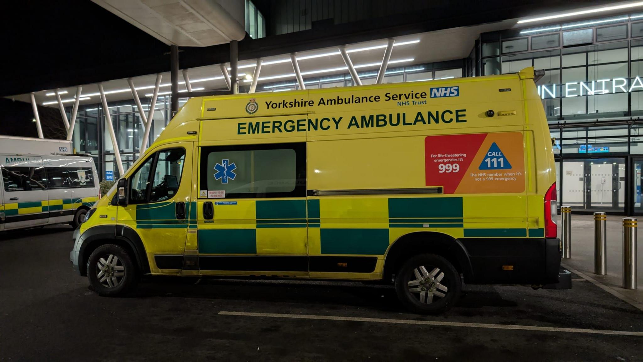 An ambulance is parked outside Hull Royal Infirmary's Accident and Emergency department. It is dark, and the hospital is lit up behind the vehicle. The ambulance is mainly yellow, with green rectangles painted along its side. There is branding of the Yorkshire Ambulance Service, and a sign with 999 written on it, as well as the NHS helpline number 101.