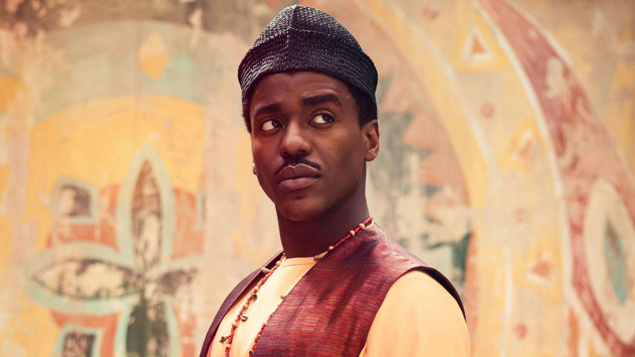 The Doctor wears a cap, orange top and red waistcoat. He stands in front of a colourful wall.