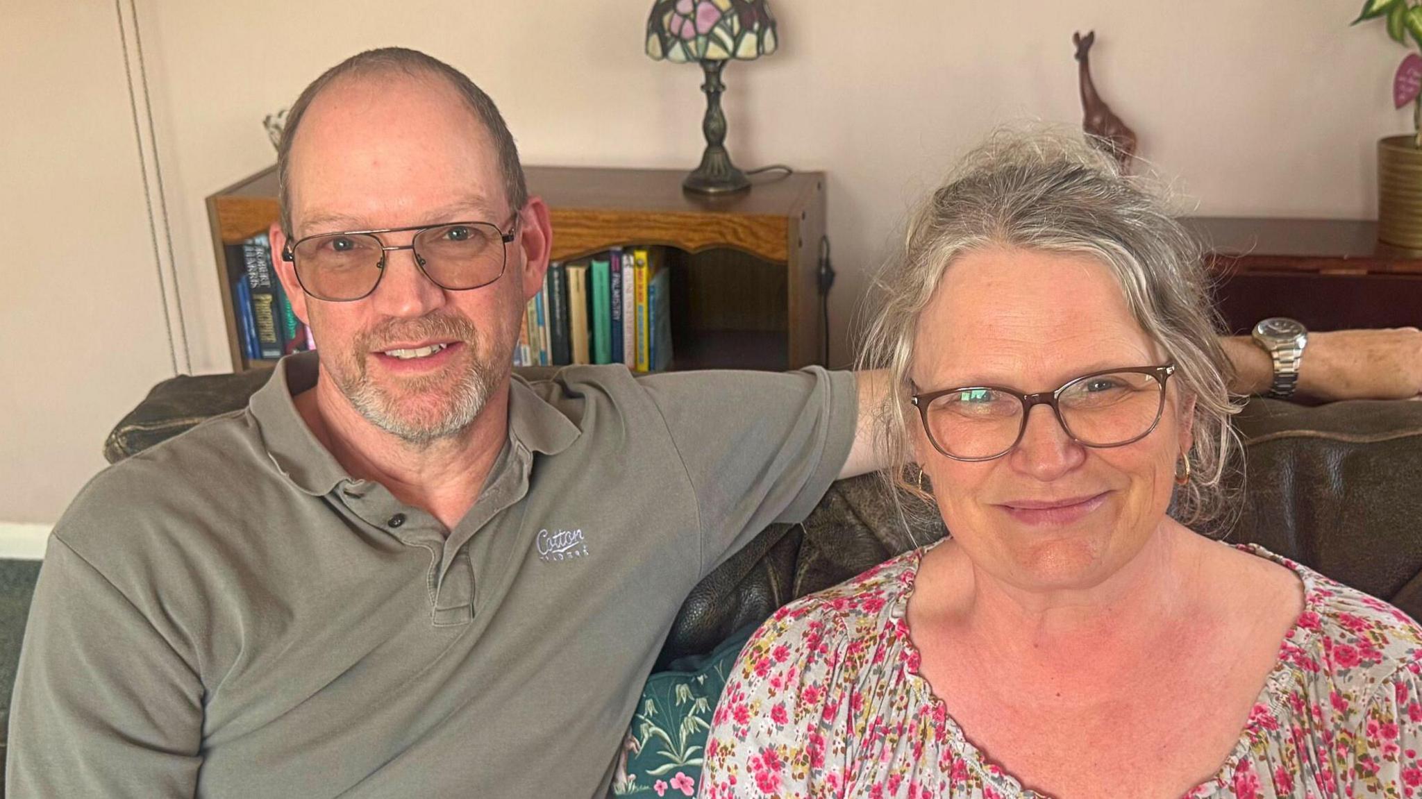 A man with glasses and a grey t-shirt puts his arm around a woman in a floral top. They sit on a sofa in a living room.