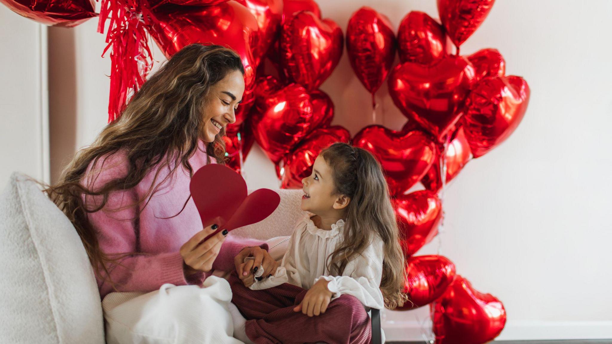 Daughter and her mother smiling and having fun with red foil heart-shaped balloons at home.