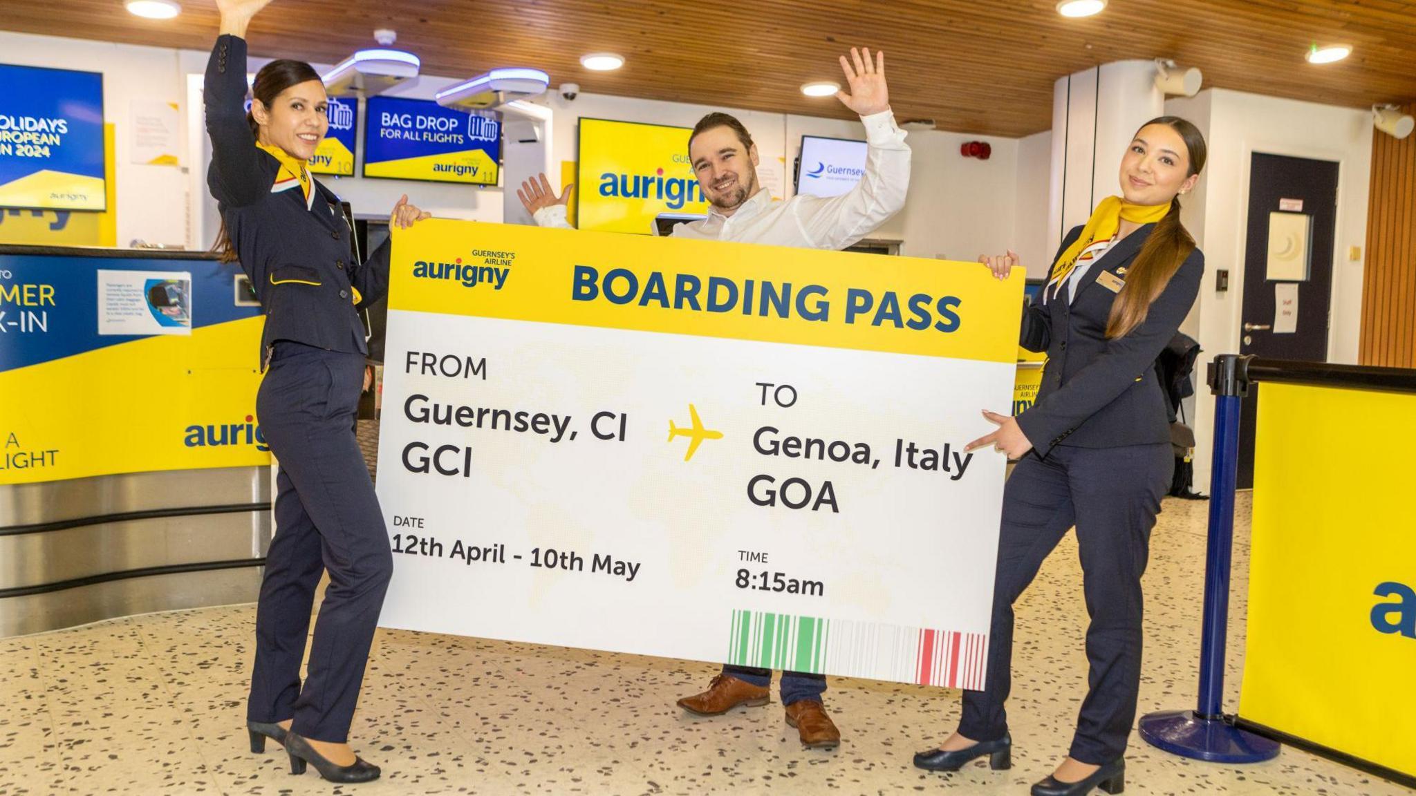 Three Aurigny staff are holding a giant boarding pass in the airport. The ticket said FROM GUERNSEY CI TO GENOA ITALY, Date 12th April - 10th May, Time 8.15am. The two women are wearing navy trouser suits with yellow detail and the man is wearing a white shirt, jeans and brown shoes. There are Aurigny yellow and blue banners and posters in the background.