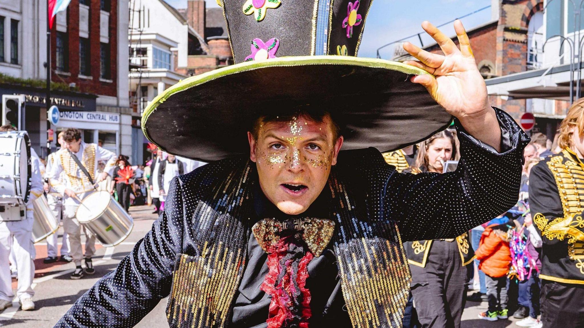 A man in an oversized top hat is staring down the barrel of the lens, as a marching band is seen playing behind him. He is in the middle of a street in Longton, Stoke-on-Trent.