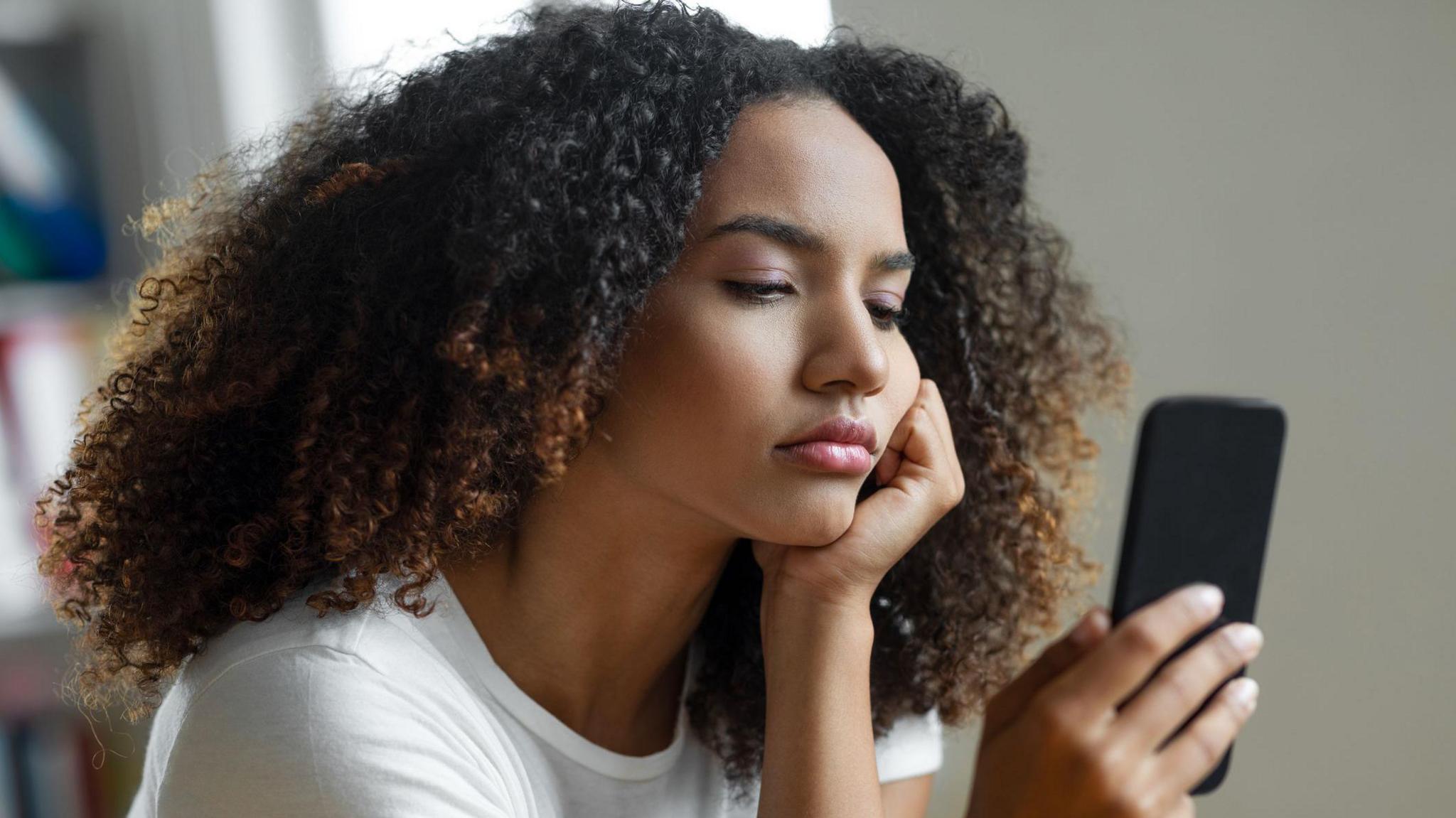 A young woman with shoulder-length hair wearing a white t-shirt, staring at her phone while looking fed up.