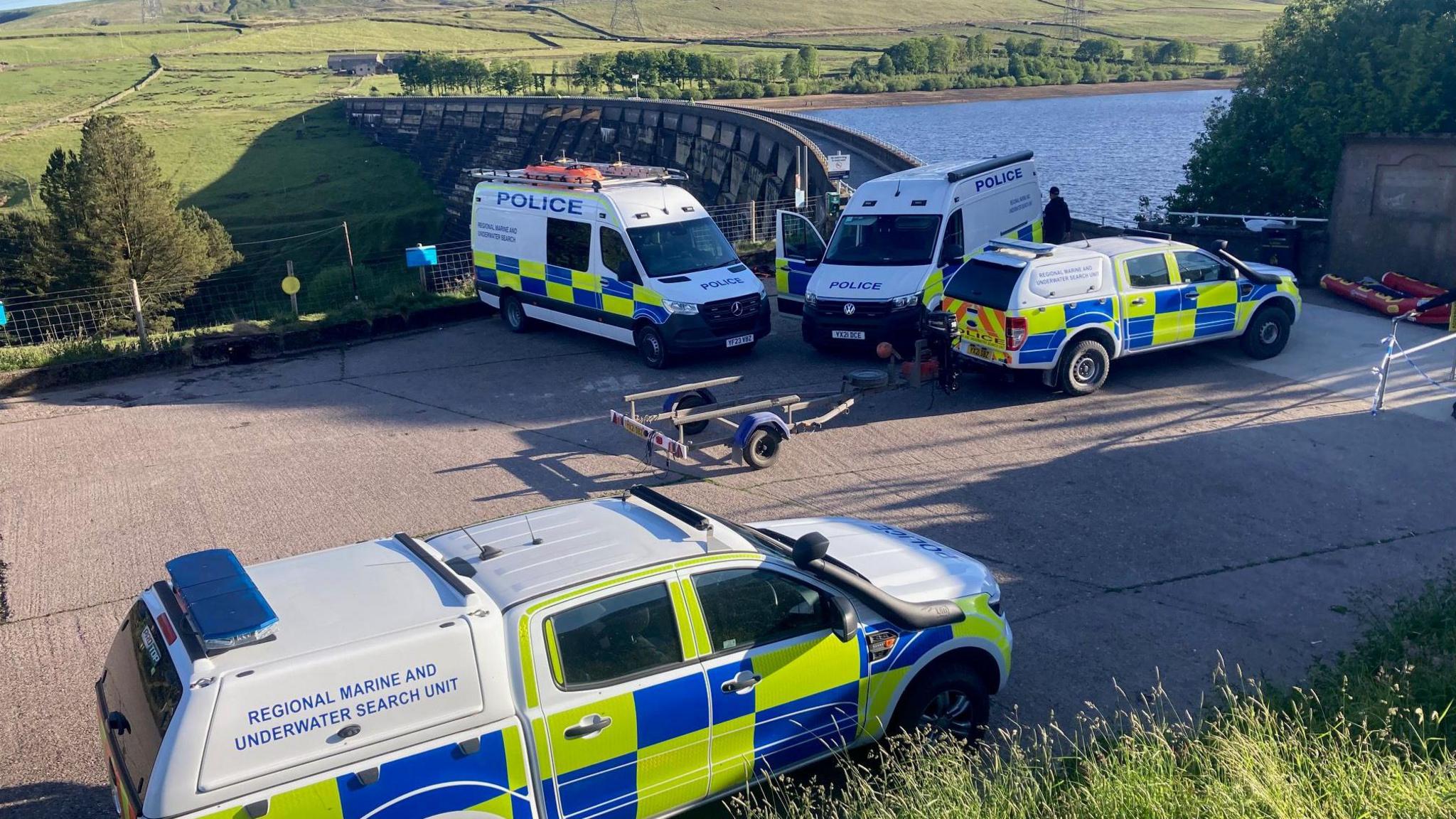Four police vans, one marked as a marine and underwater search unit, parked outside a reservoir