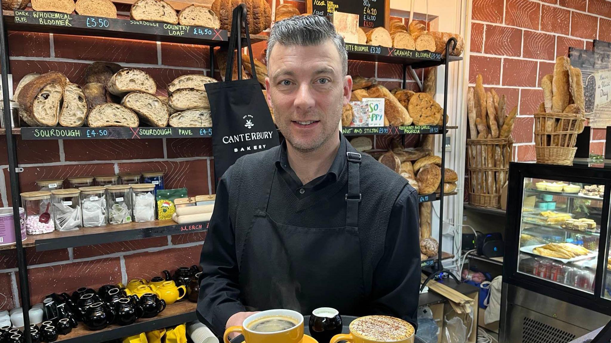 Neil Terry, Front of House Manager at The Canterbury Bakery, stands in front of a shelf of bread loaves holding a tray of coffees