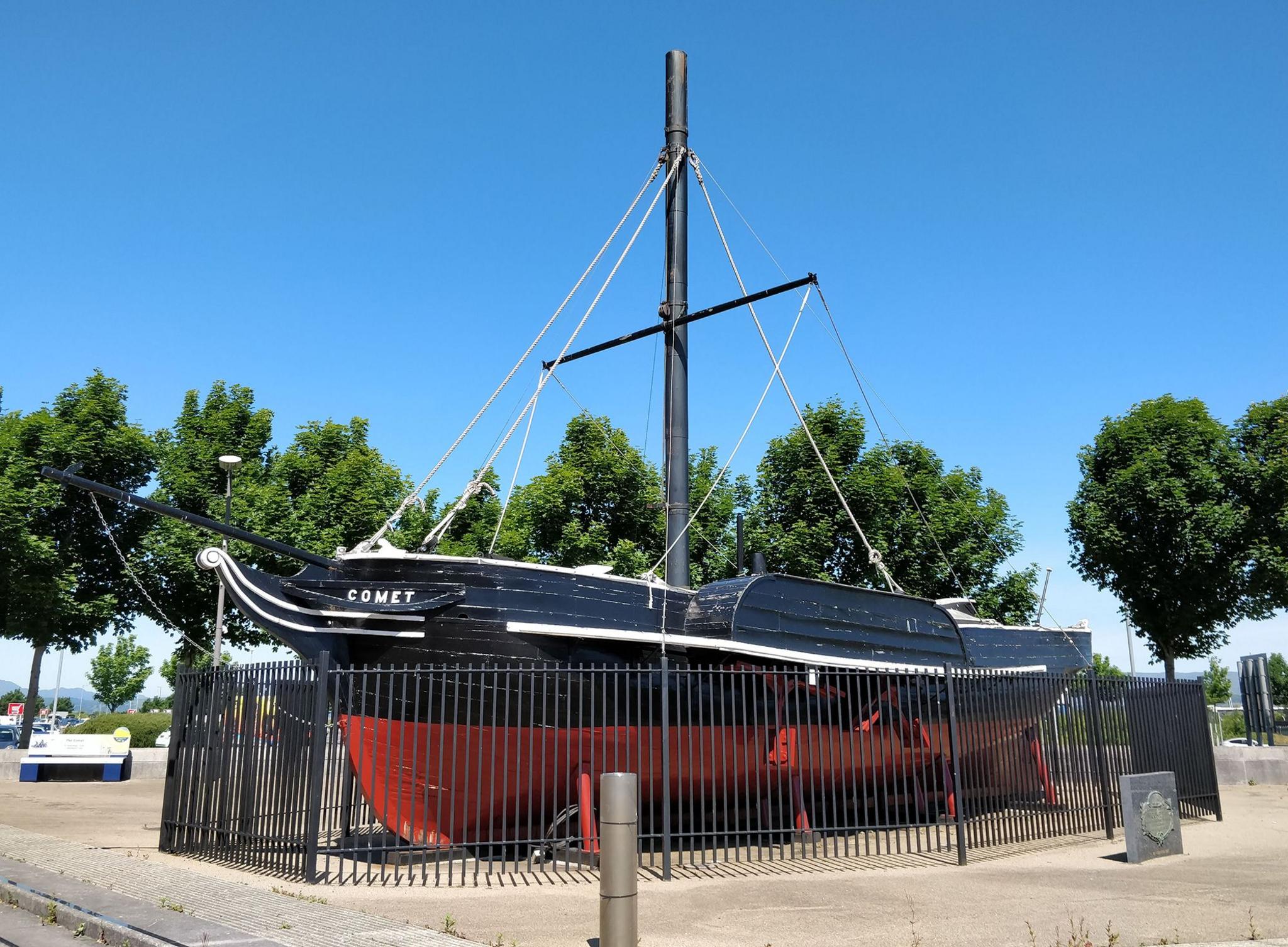 A black and red wooden ship with paddles at the side and a thin funnel. The ship is surrounded by black railings. There are trees in the background.