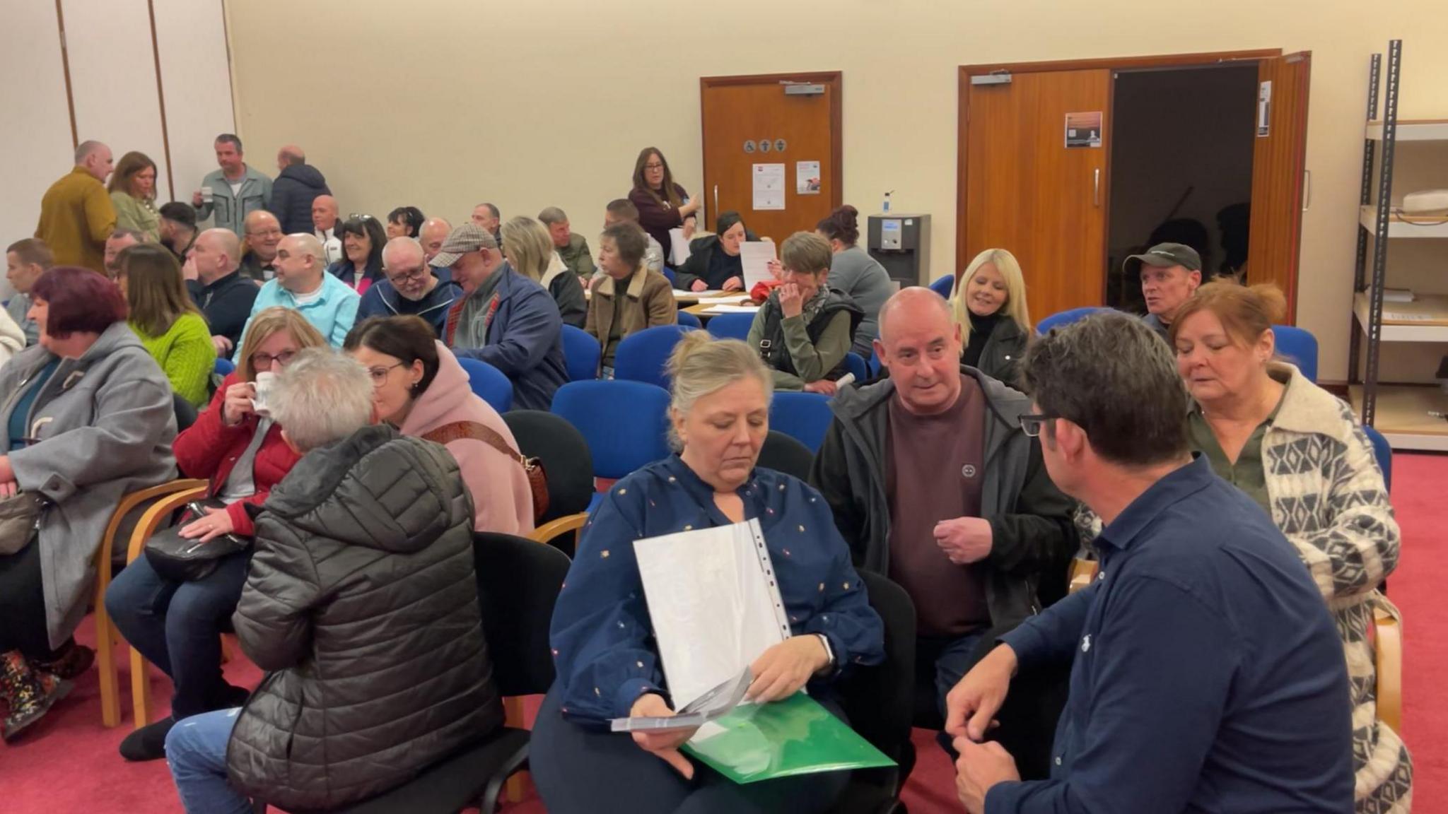 A group of former Royal Stafford workers sat chatting in a large room. There are men and women on chairs, some with pieces of paper while others chat.