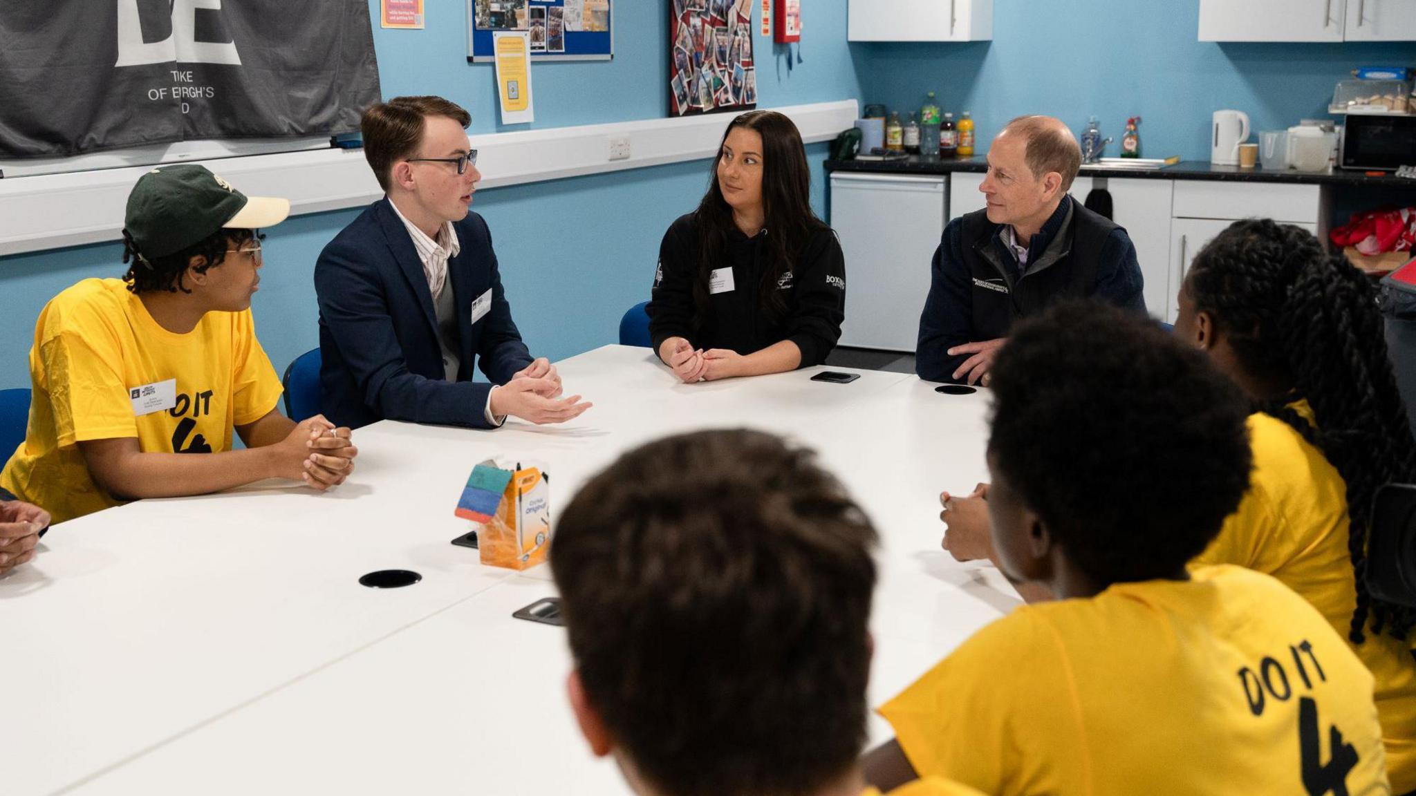 Young people wearing T-shirts with around a table with the Duke of Edinburgh and two other adults, chatting.