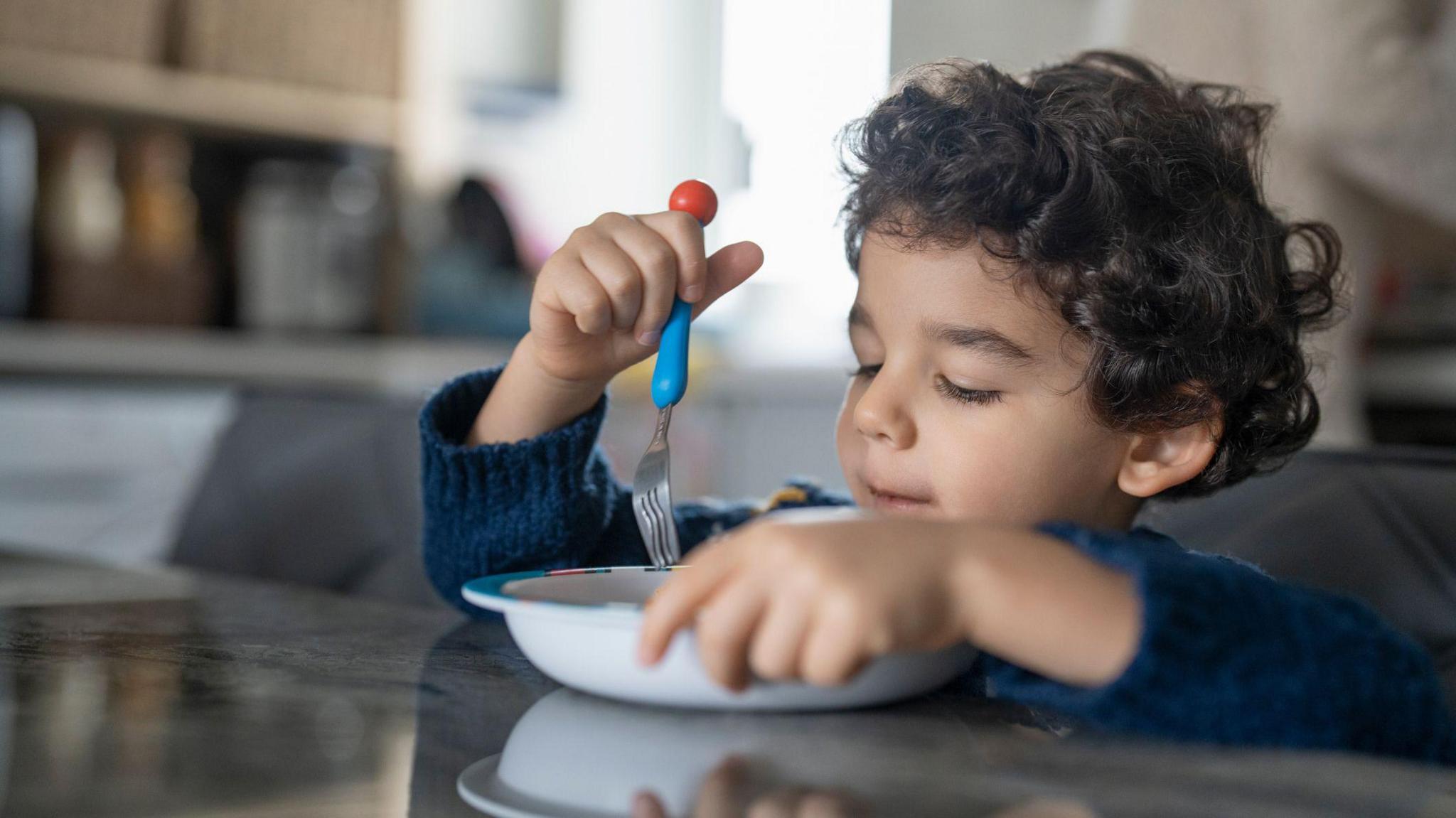 A child with brown curly hair wearing a dark blue jumper eating with from a white bowl with a fork. 