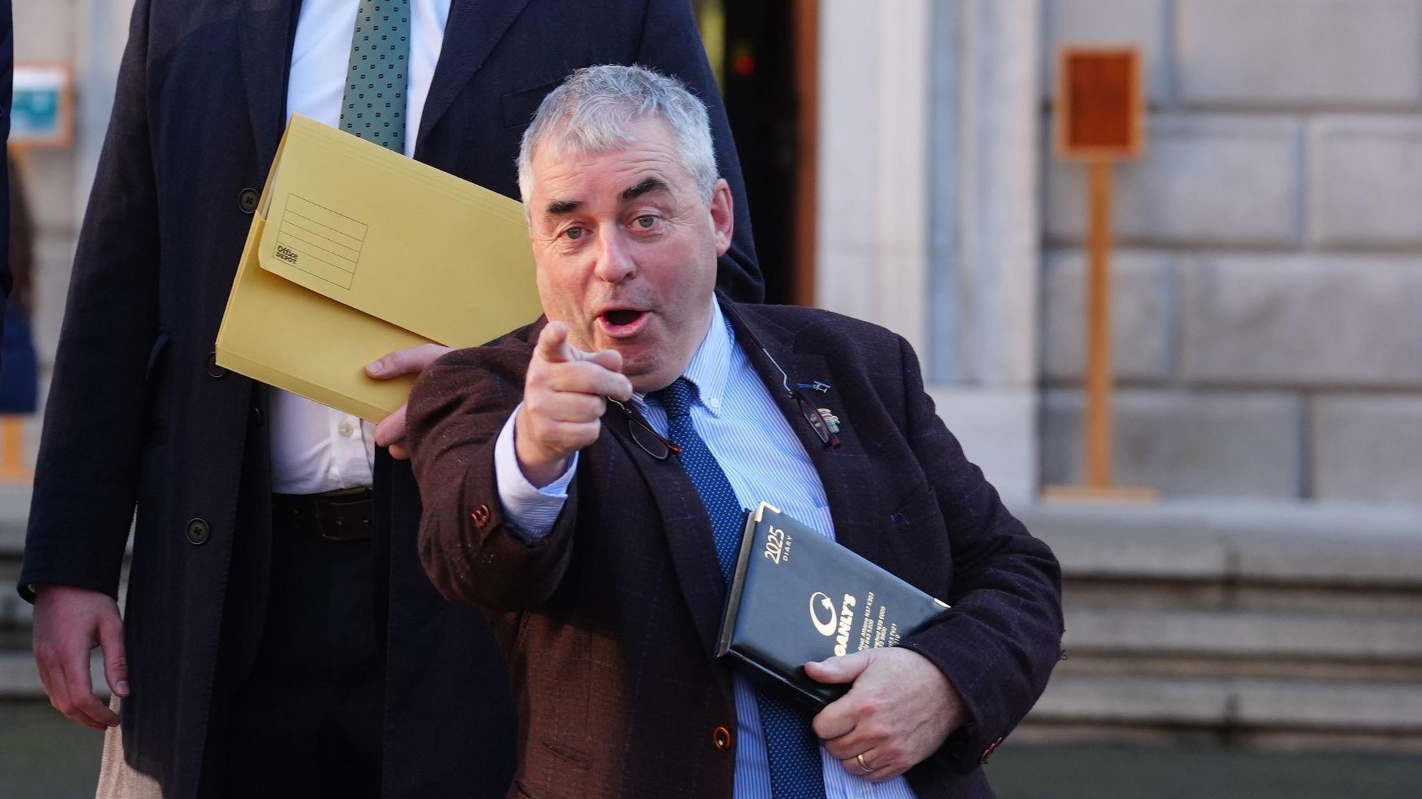 Kevin Boxer Moran. He has short grey hair and blue eyes, wearing a dark blazer, light blue shirt and dark blue tie. He is pointing at the camera in a jokey way and holding a leather diary outside Leinster House in Dublin.