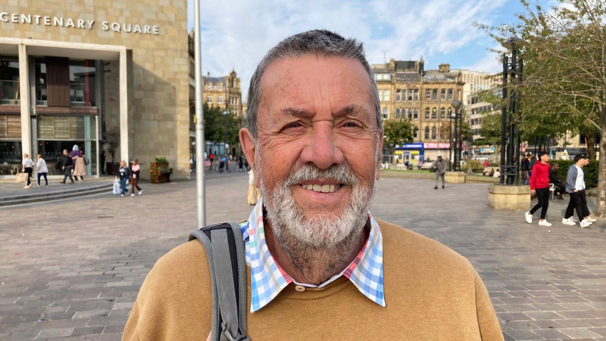 A white man with grey hair and a grey beard looks at the camera. He is wearing a mustard coloured jumper with the collar of a blue and white checked shirt visible at the neck line. He has a bag slung over his right shoulder