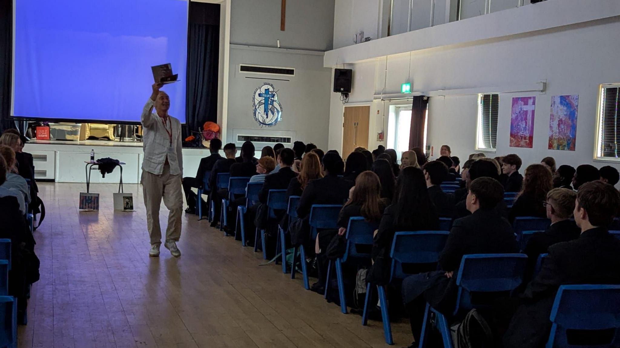 Mike Garry in hall of school children holds a book in the air - a blue screen can be seen in the background