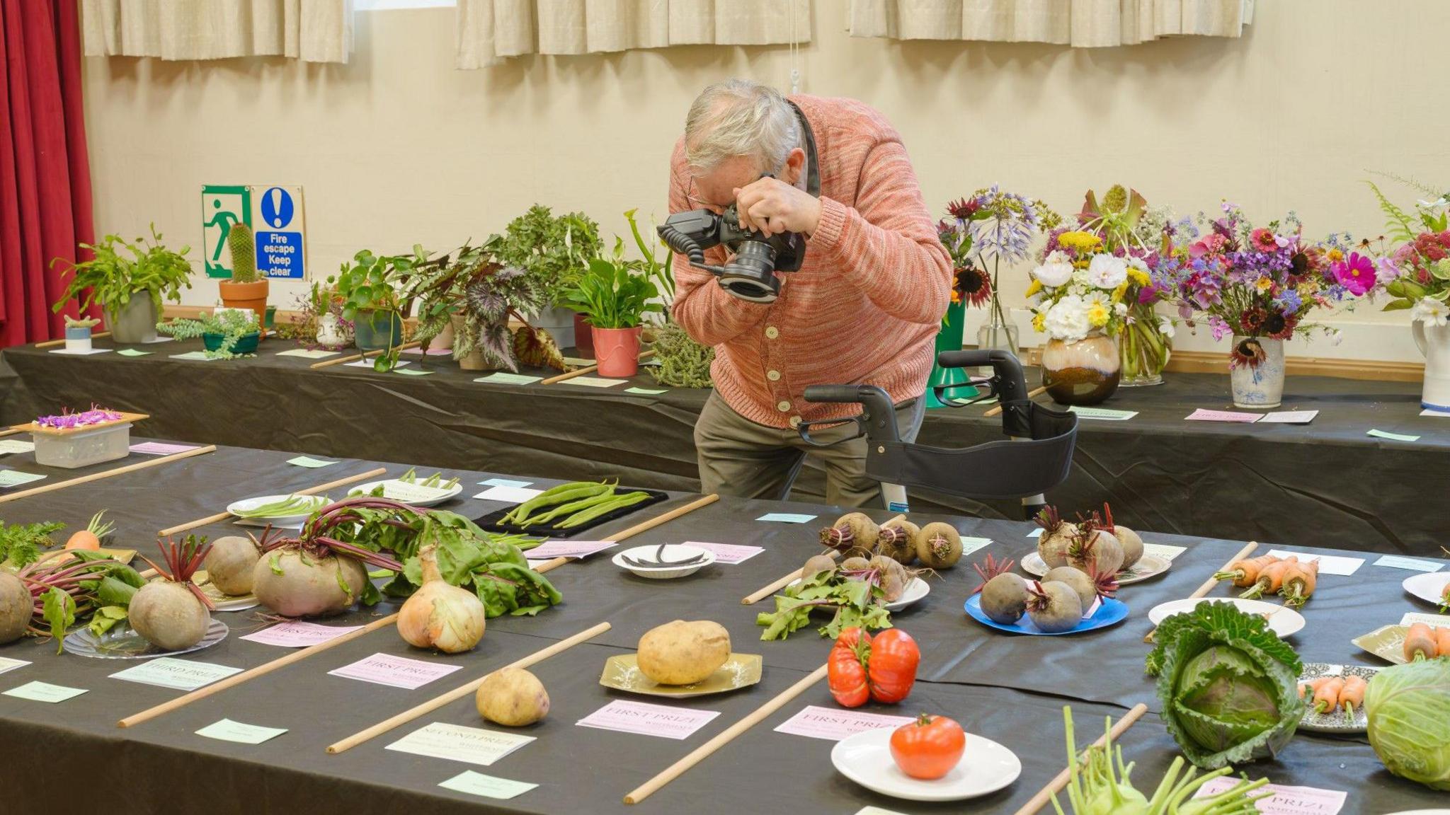 A man with a camera leaning over a table of vegetable entries at a village show