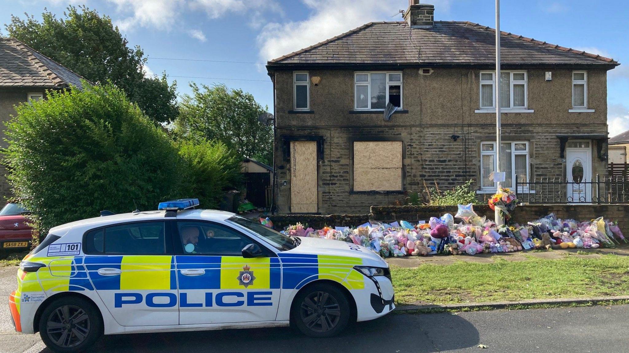 A police car parked outside the burnt out house which also has flowers and cuddly toys laid against a garden wall 