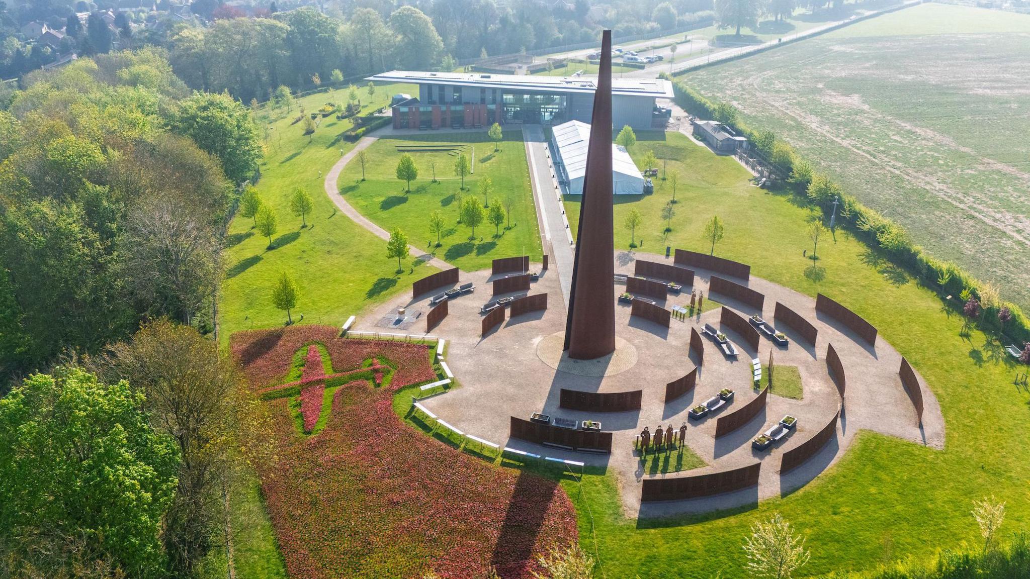 The poppy display at the International Bomber Command Centre which was installed to mark the 80th anniversaries of VE and VJ Day. There is a tall spire and memorial walls surrounded by grass and trees. There are red ceramic poppies arranged in the silhouette of a Lancaster Bomber.