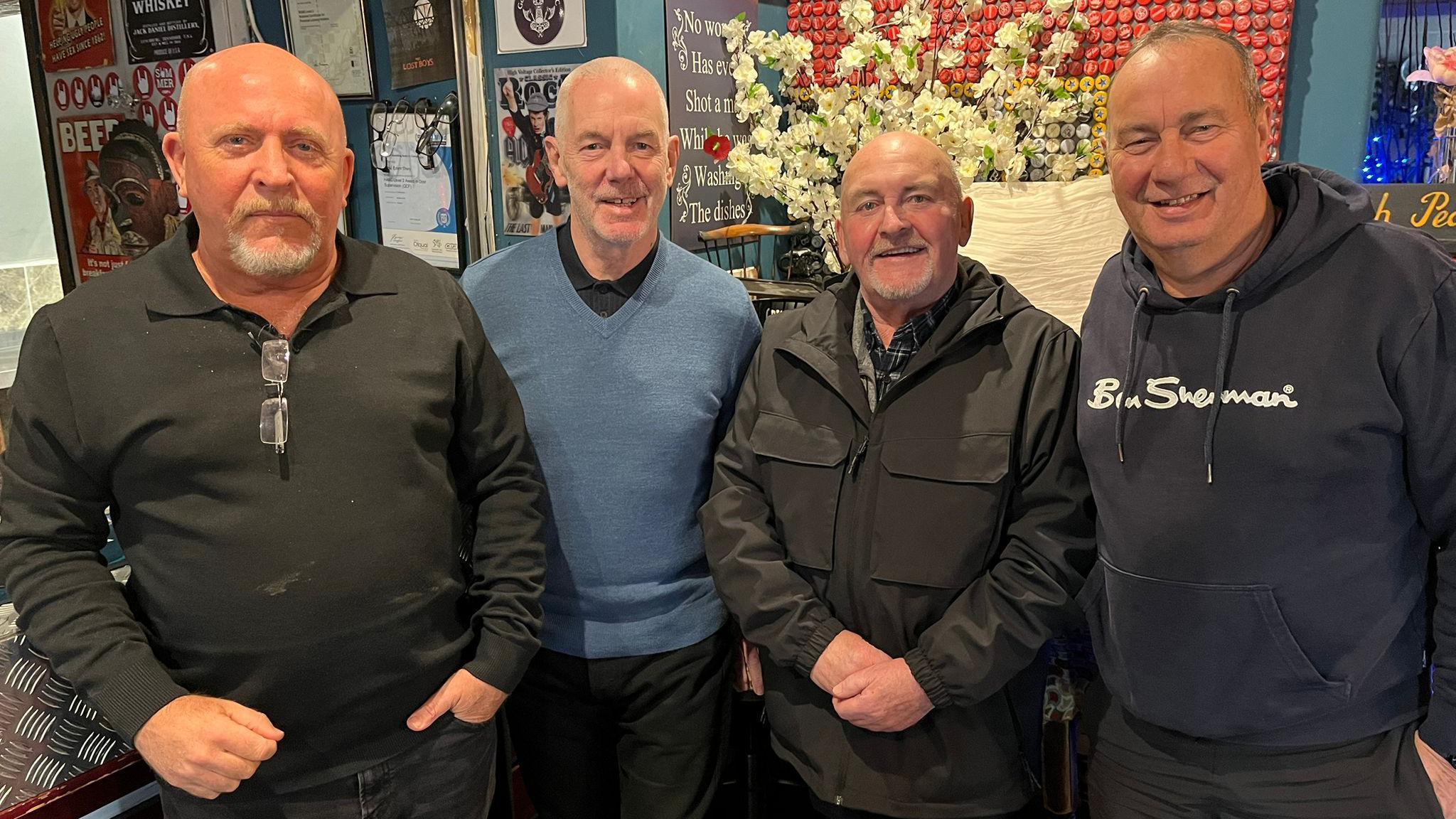 Four men in a line all wearing dark colours. These are left to right