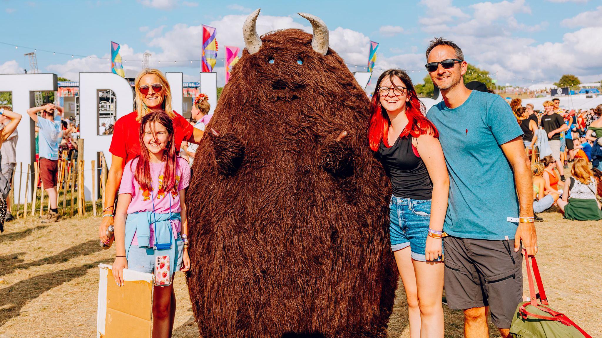 The festival mascot the Truck Monster, which is a fluffy bull-looking creature with horns, is taking picture with a family of four. It is sunny. Other festival-goers can be seen behind them.