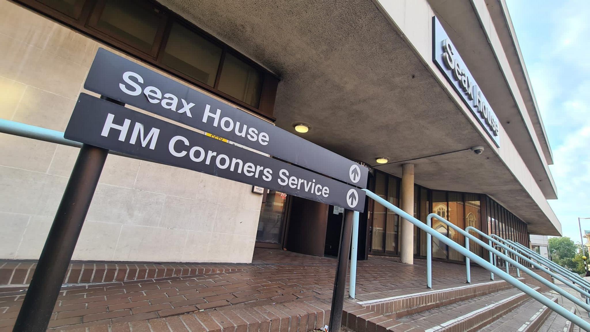 The outside of Seax House in Chelmsford, where the Essex Coroner's Service is based. It is a modern concrete building with three storeys with wide paved steps and railings leading to the entrance. A sign says Seax House HM Coroners Service.