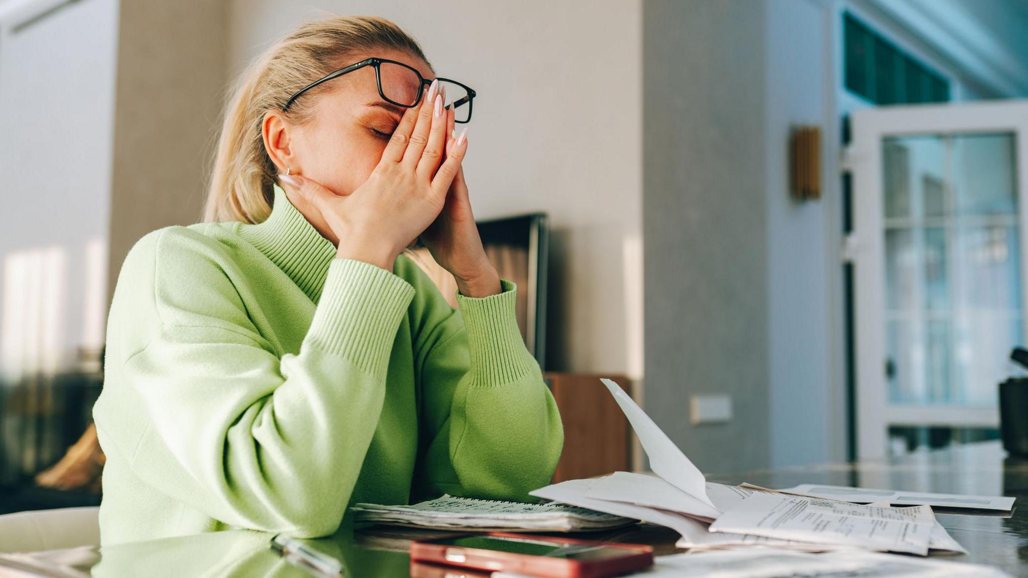 Woman sits at a table at home with her head in her hands, with paperwork in front of her.