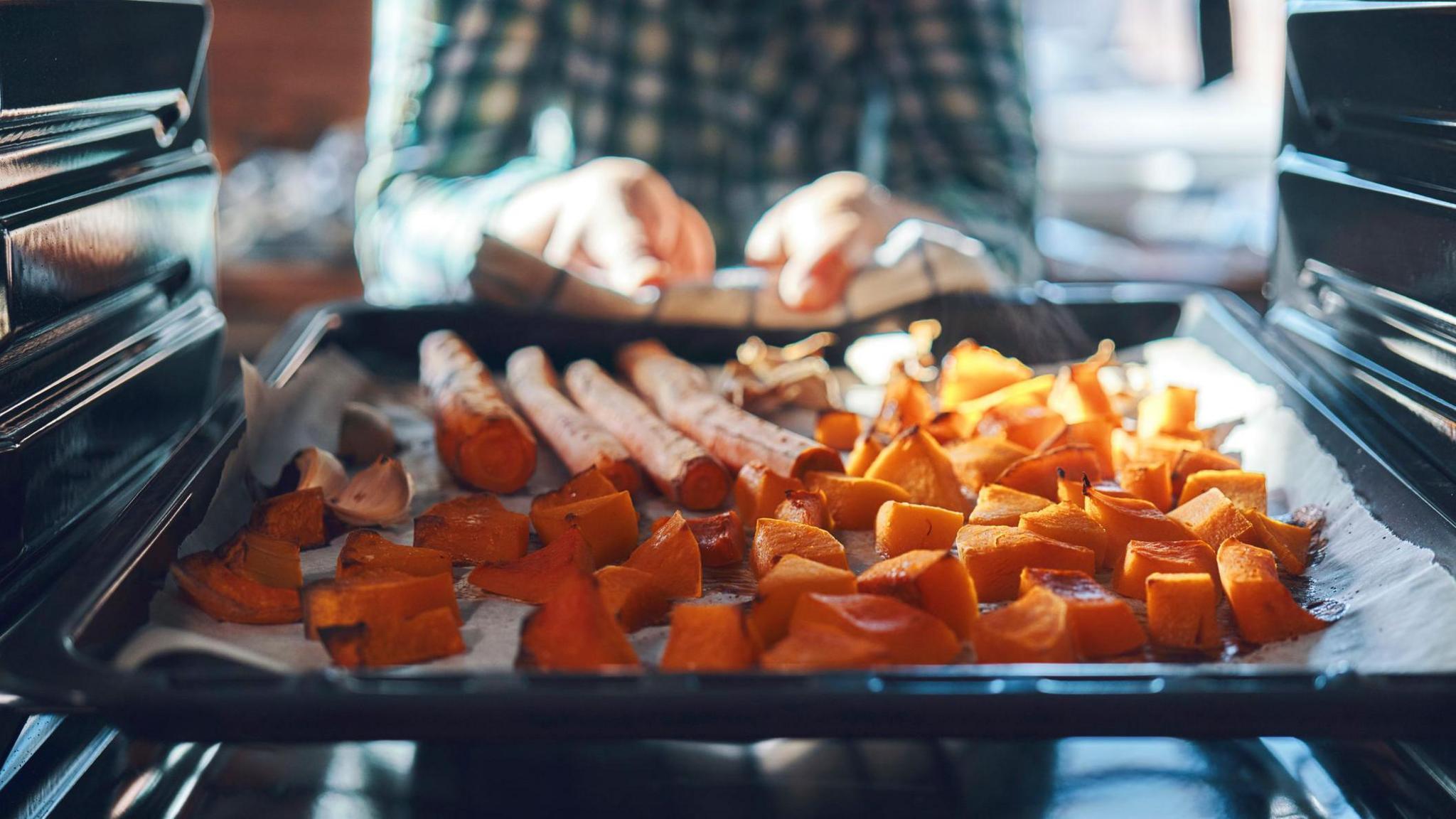 A stock image of a man pulling a tray of roast pumpkin out of the oven using a tea towel.