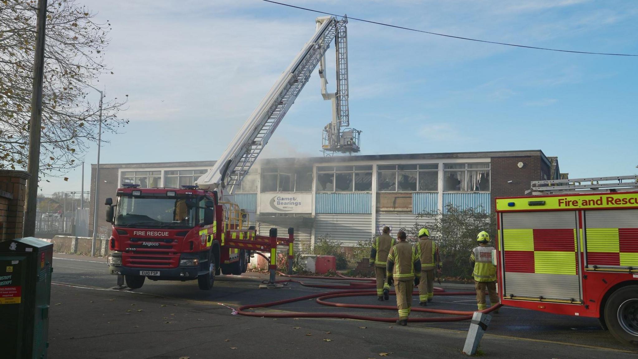 Two fire engines are outside the derelict building, one has an aerial ladder platform. There are four firefighters in protective clothing walking past one of the fire engines towards the building.