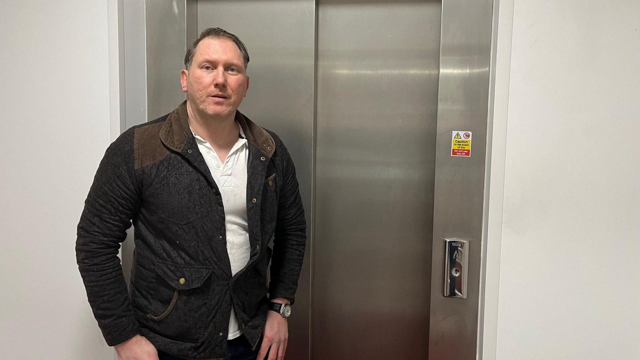 Stephen Dean standing in front of one of the lifts which is due to be replaced. He has dark hair brushed to one side and stubble. He is wearing a dark coat and white top. 