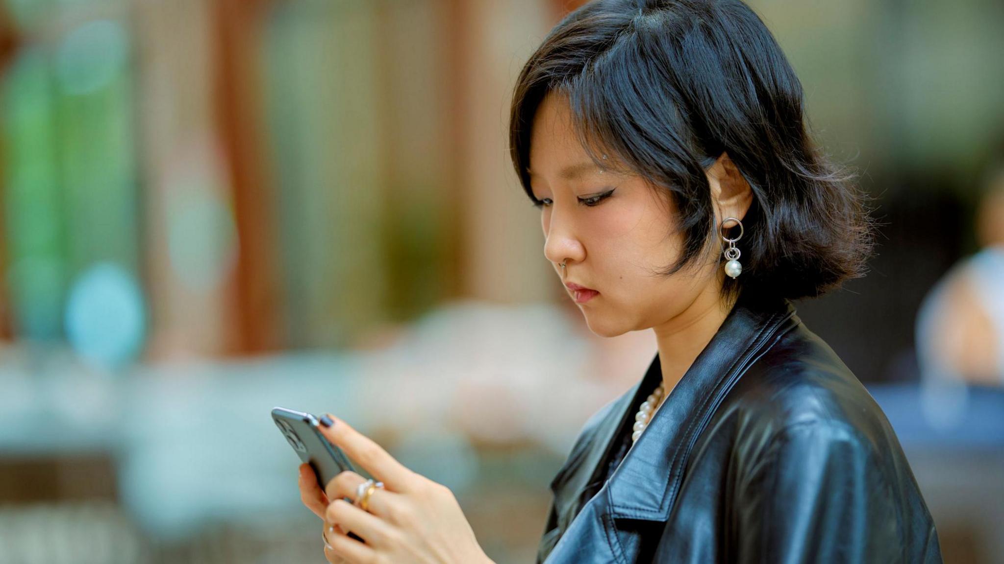 Young woman in a leather jacket using a smartphone in an outdoor setting. Shanghai, China - stock photo