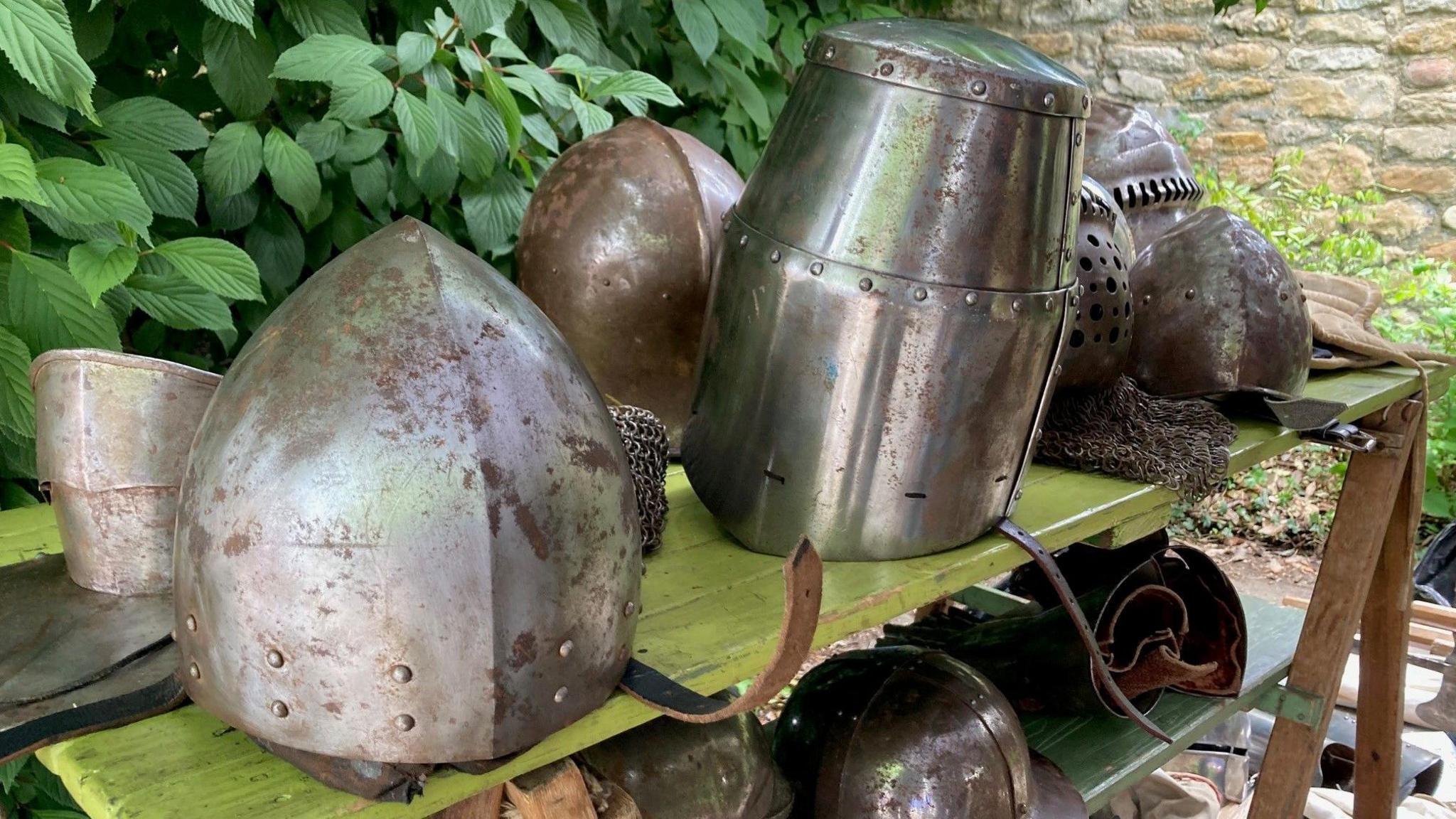 Various metal helmets and pieces of chain mail sit on a wooden table next to some shrubs.