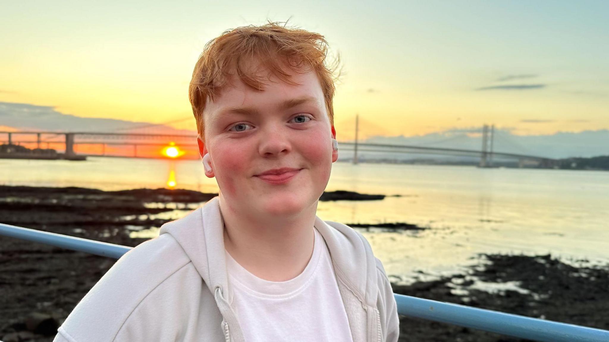 Mr Horne is smiling at the camera while standing in front of a large body of water during a sunset. He has ginger hair and is wearing a white top, light jackets and airpods in his ears. The sky behind him is cast in an orange and yellow glow.