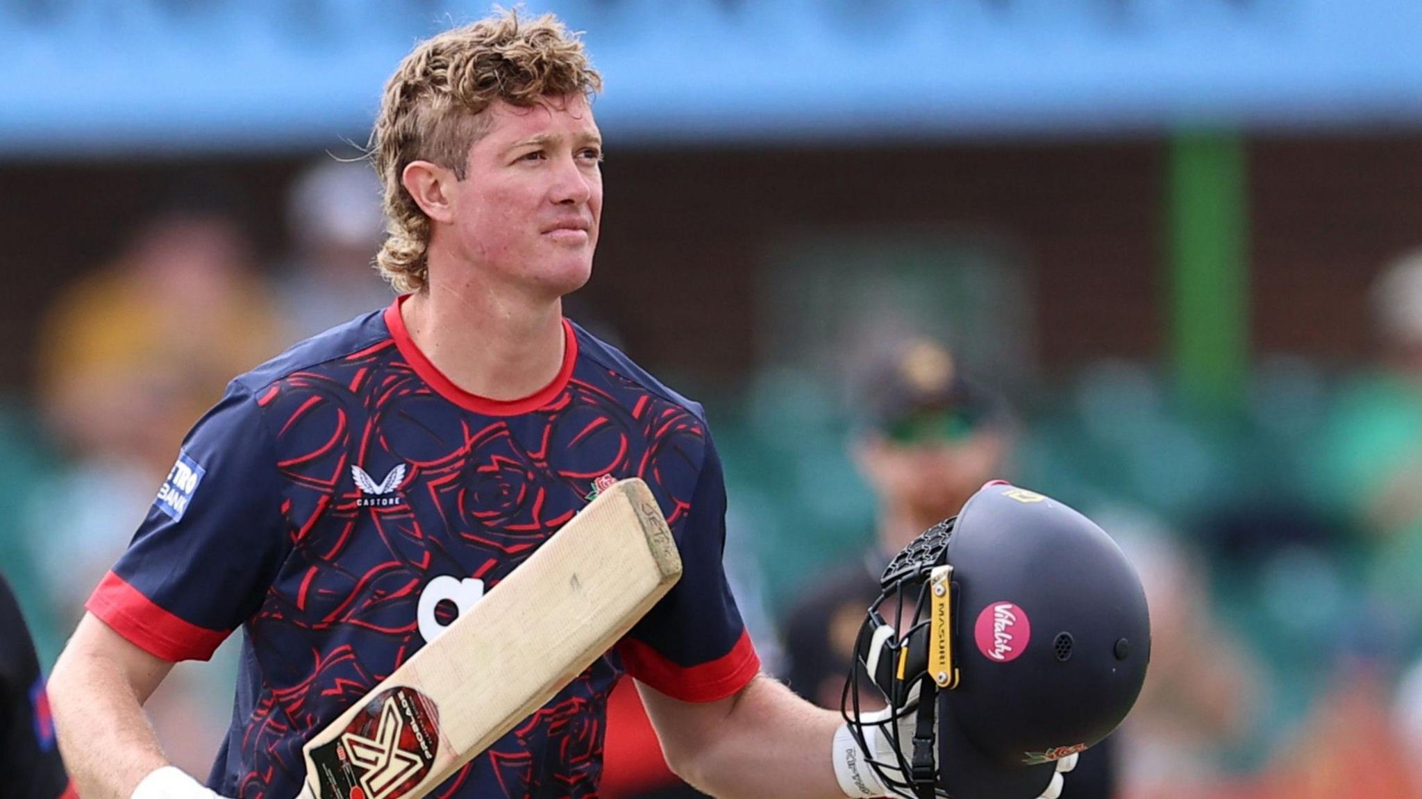 Keaton Jennings holds his helmet and bat wearing a blue and red Lancashire kit