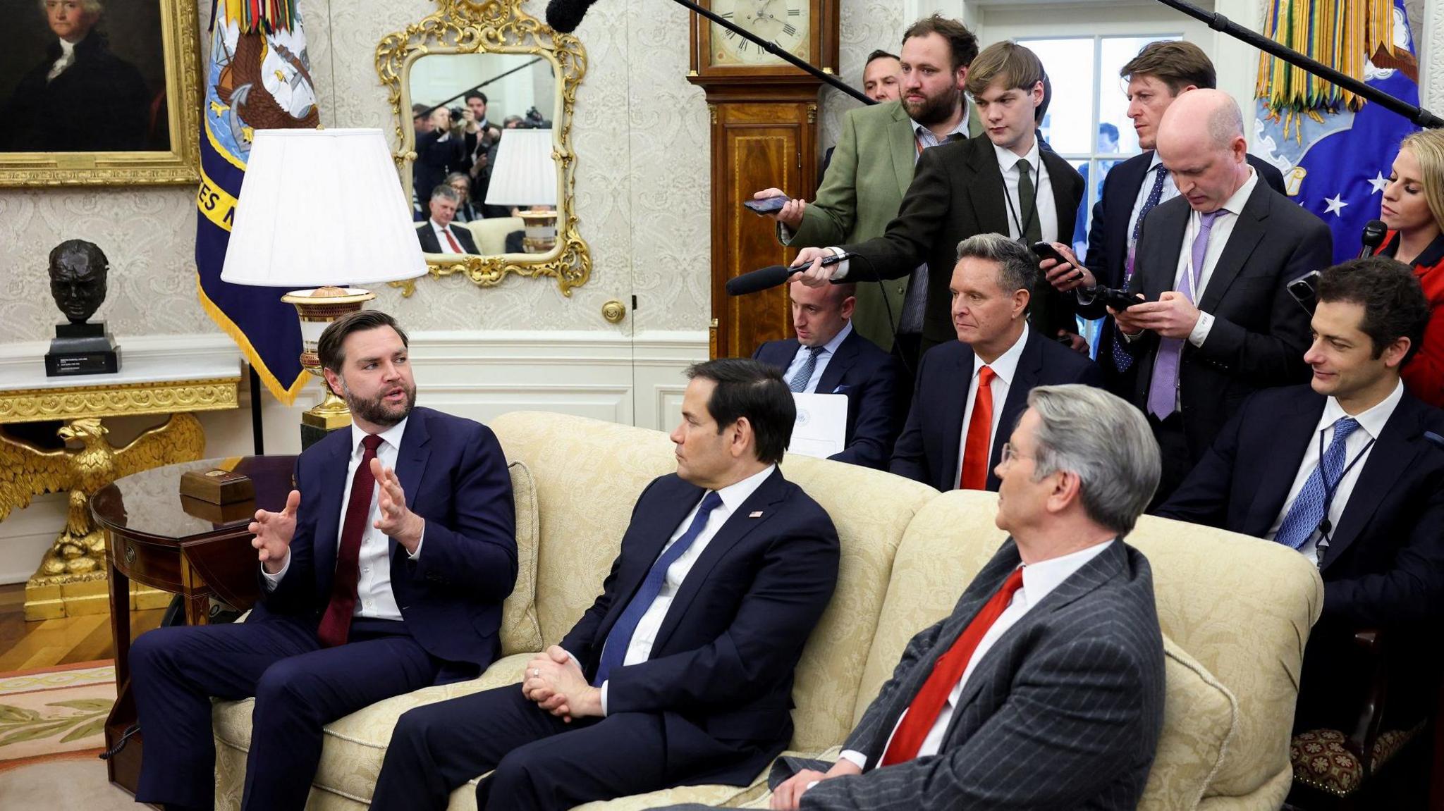 US Vice President JD Vance, sitting on a sofa in the Oval Office, speaks to a group of reporters behind him holding microphones. 