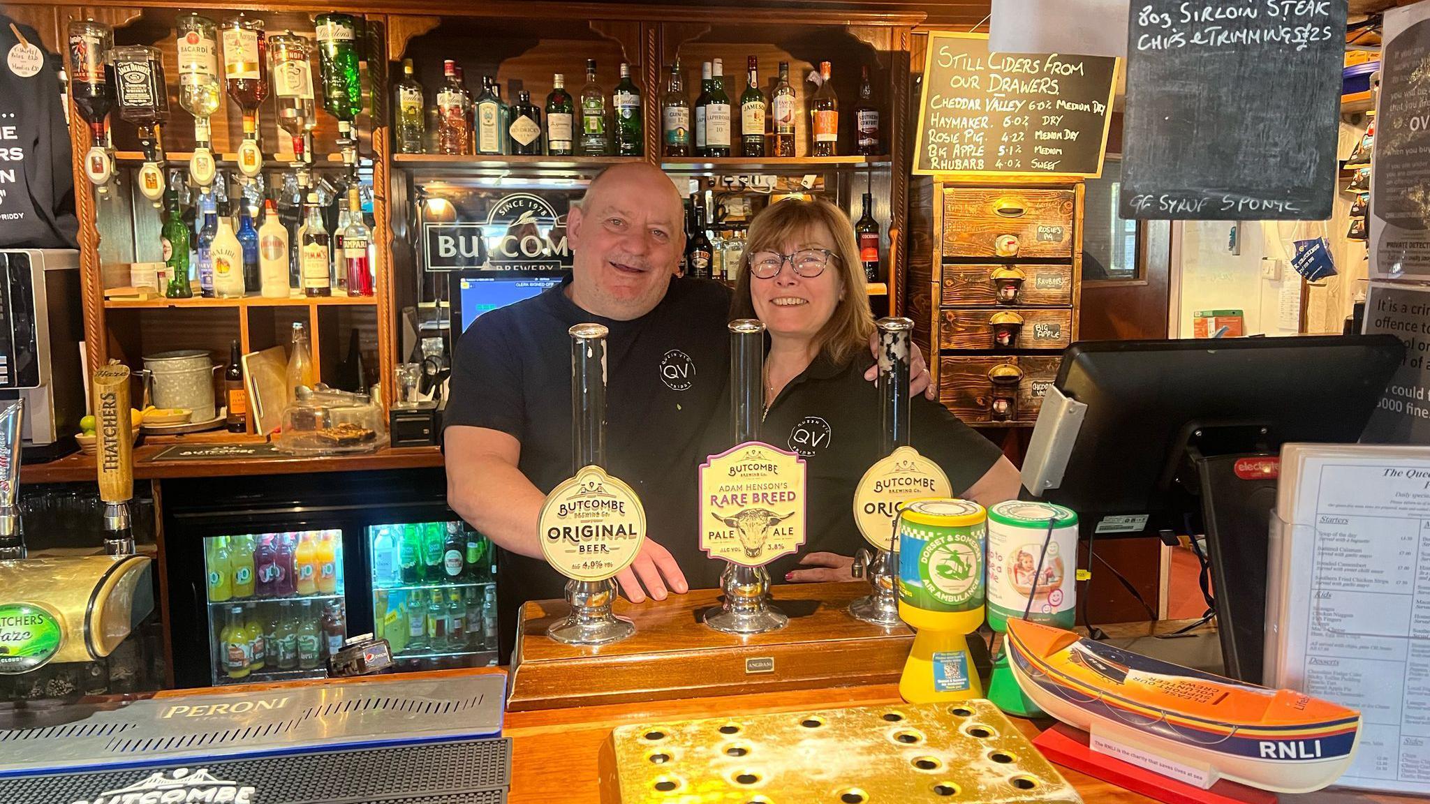 Two pub landlords in matching black shirts smile as they stand behind the bar at a pub.