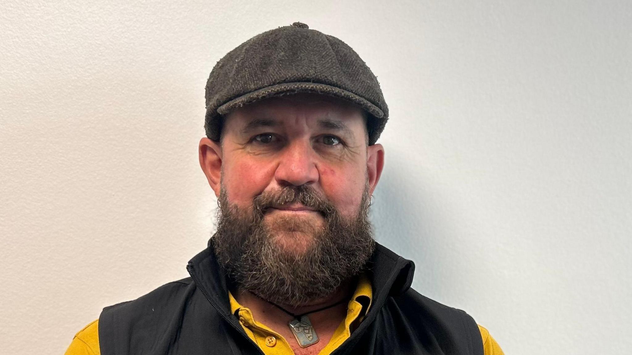 Jonny Yaxley is standing against a white wall. He is wearing a yellow polo shirt, black gilet and flat cap.