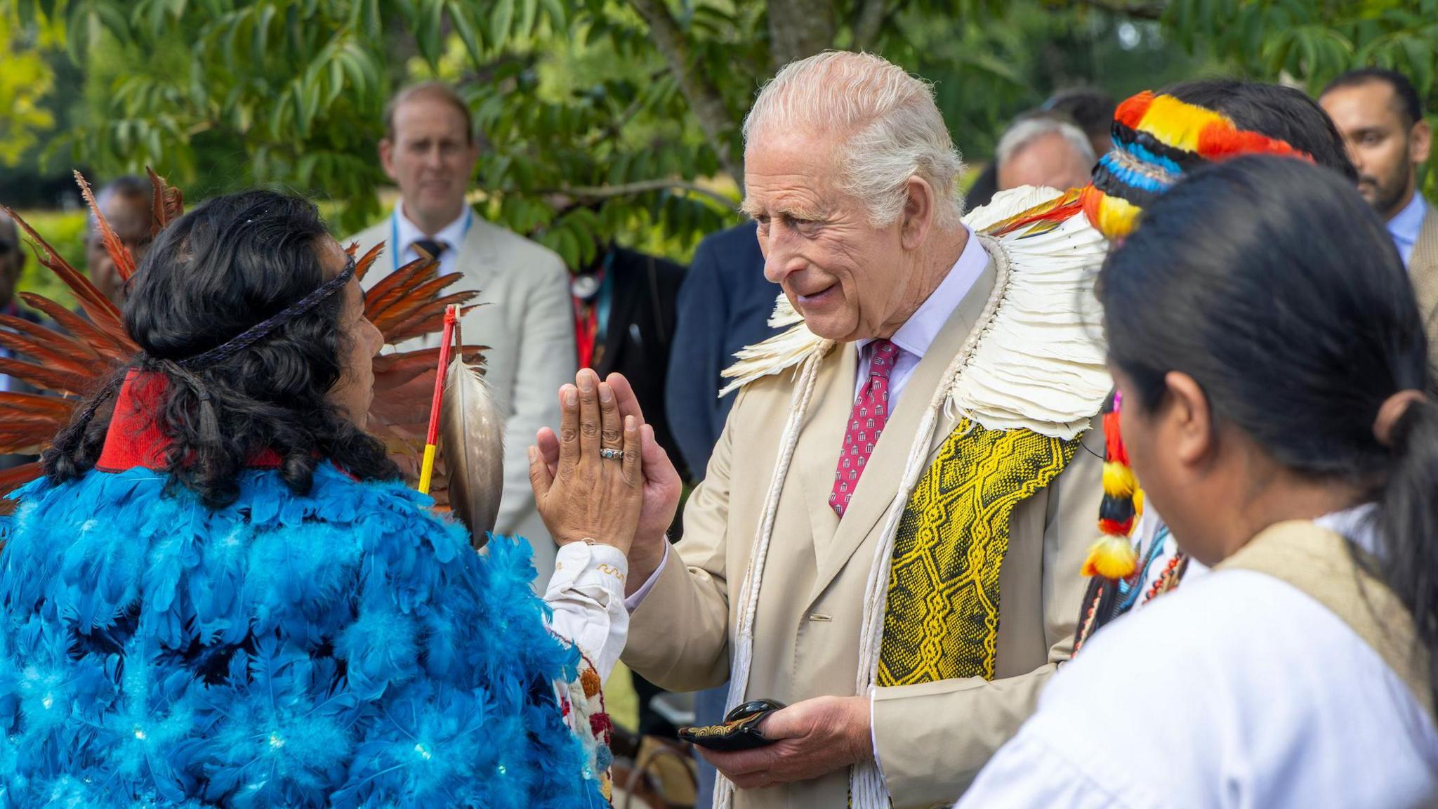 King Charles, in a suit and tie, meeting an Indigenous leader wearing a blue cloak made from feathers, at an event in Highgrove in July 2025