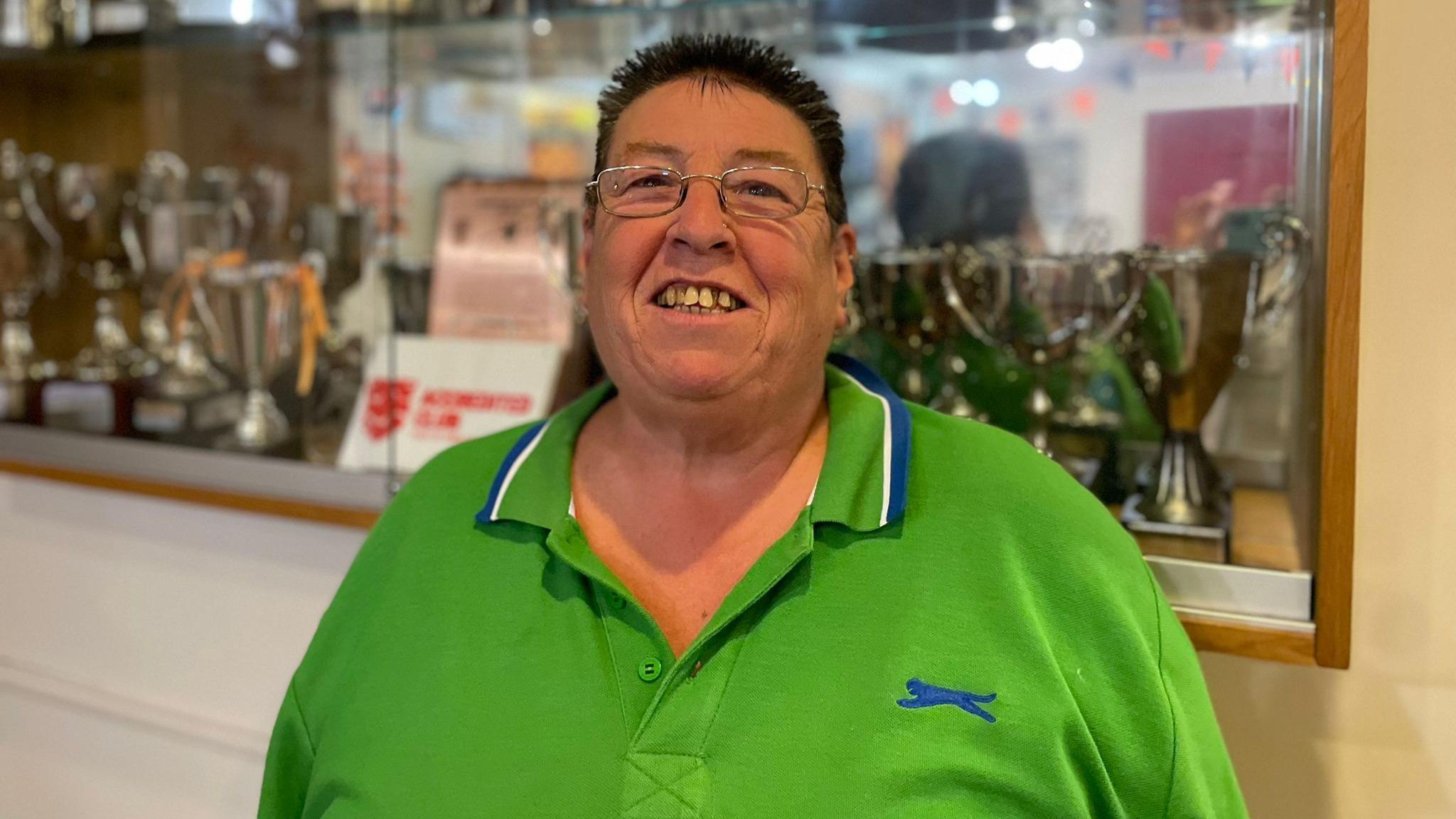 Linda is stood infront of a trophy cabinet. She's wearing a green polo shirt and is smiling at the camera.