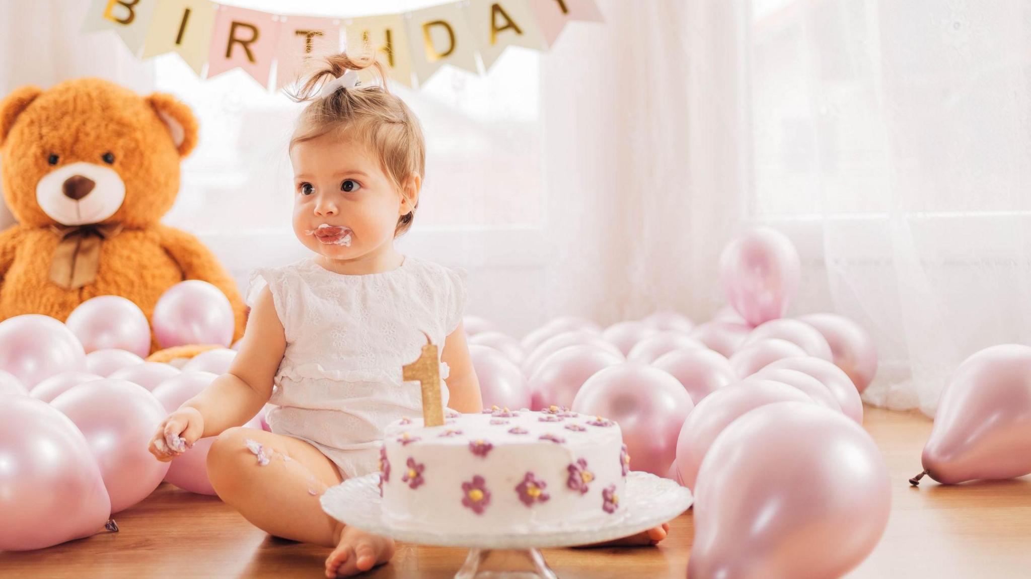 A baby girl sits cross-legged on the floor in front of her first birthday cake decorated with purple flowers and a one-shaped candle. She's surrounded by pink balloons, a giant teddy bears sits in the background.  