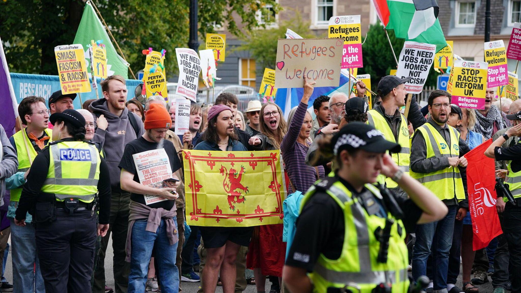 A group of people hold signs which say things like "refugees welcome" and "smash fascism and racism". Police flank the group