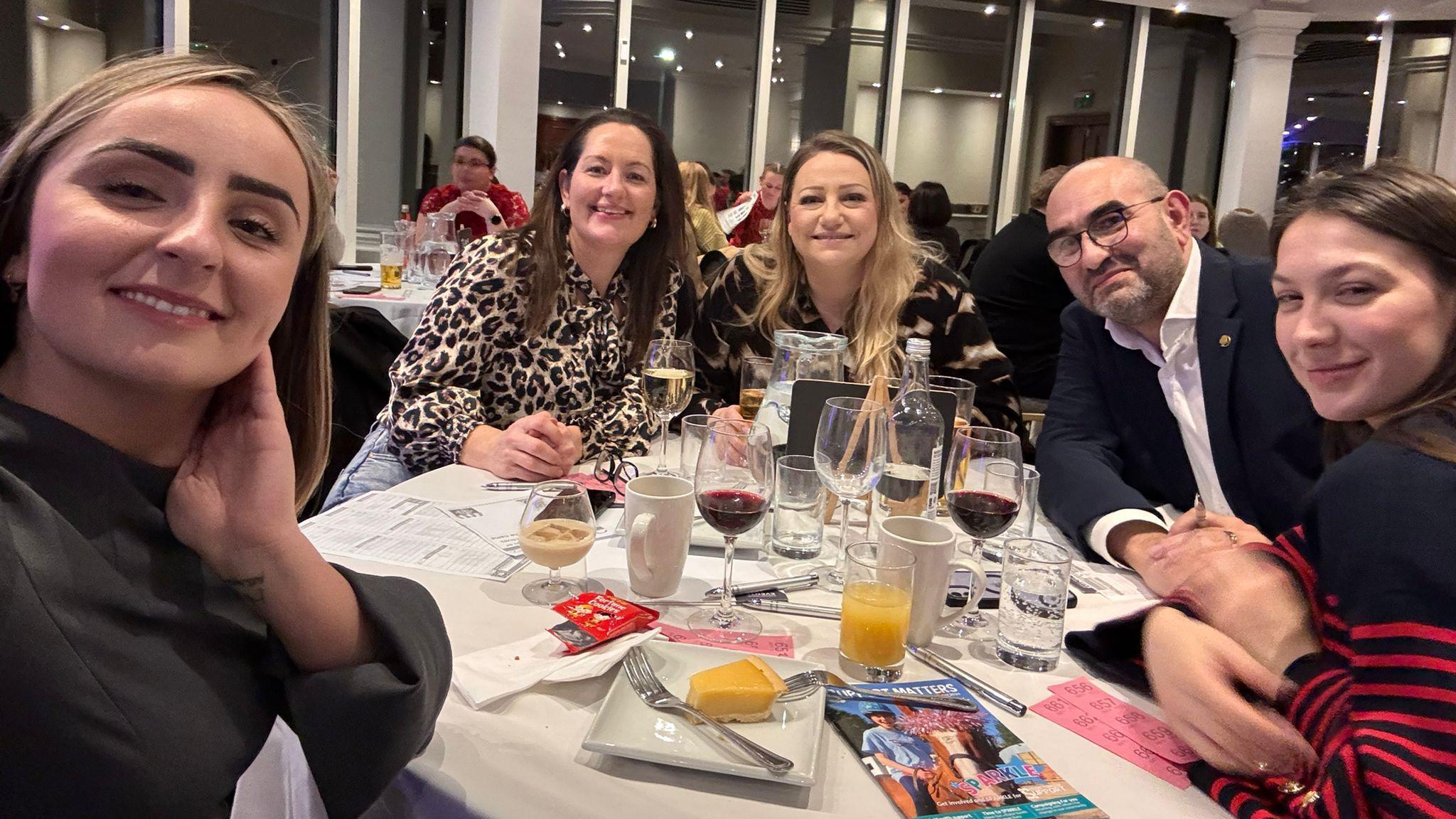 A group of people at a quiz event pose for a selfie as they sit around a round table. On the table in front of them are various glasses, some containing orange juice and red wine and plates of dessert. The table has a white cover on it