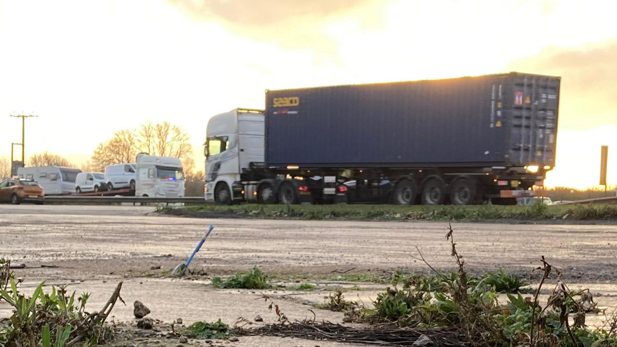 A view across a layby towards traffic on the A1, including a large blue lorry with a white cab, and a white car transported with small white vans on the back. 