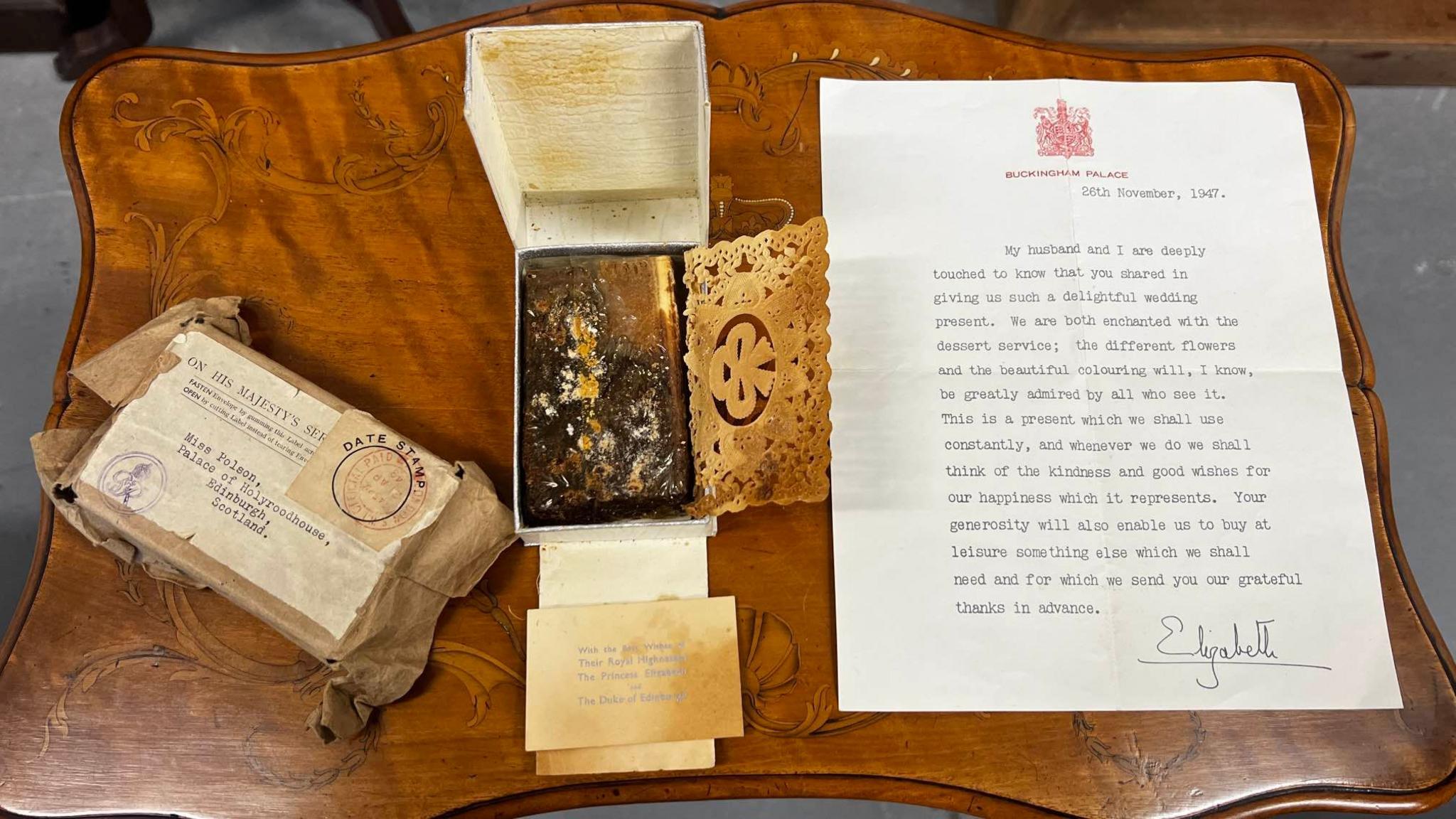 The cake, which is a fruit cake that looks mouldy, next to a sealed presentation box and a letter from the Queen. It is all laid out on a brown wooden table.