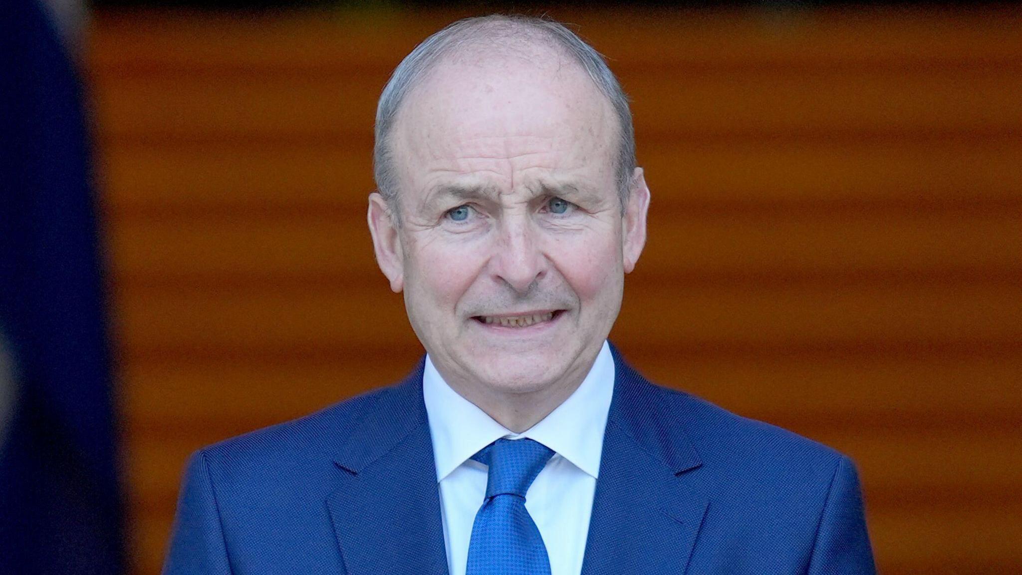 Taoiseach Micheál Martin, pictured from the chest up. He has balding short grey hair and he is wearing a dark blue suit, blue tie and white shirt. 