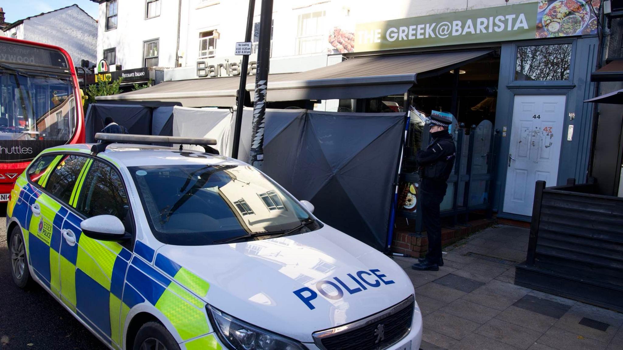 A police officer stands in front of black screens and a police car at the Barista venue, which has a black canopy and green banner across its entrance. It is based on a busy city street scene.