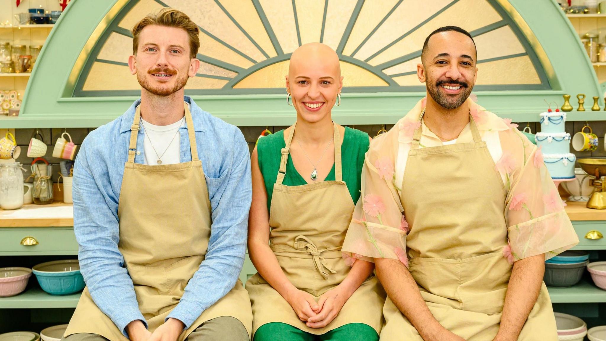 Tom in a blue shirt and beige apron, Jasmine in a green jumsuit and beige apron and Aaron in a flowery shirt and beige apron sitting in the Bake Off tent.