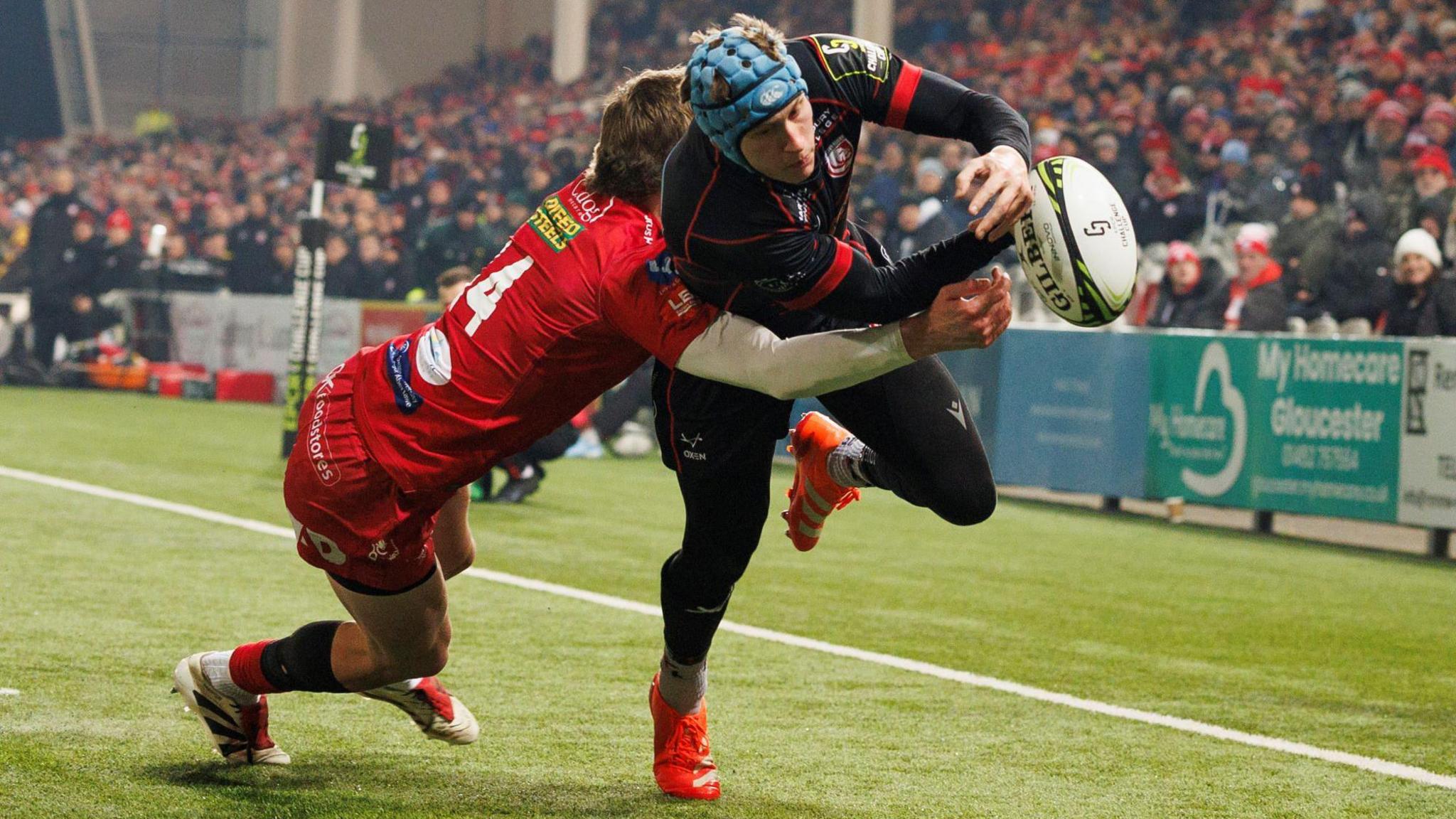 Gloucester's Josh Hathaway leaps towards the try line as he is tackled by a Scarlets player during a match at Kingsholm. The ball has been knocked out of Hathaway's hands during the challenge. A packed main stand is visible in the background