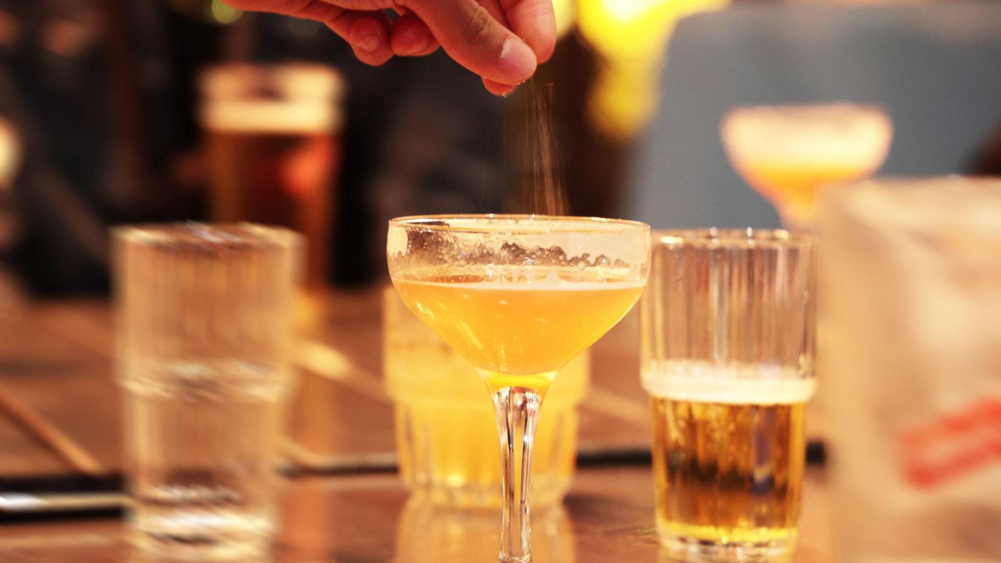 An artistic image showing a number of drinks on a bar and a hand sprinkling some unidentified powder into a cocktail glass in the foreground