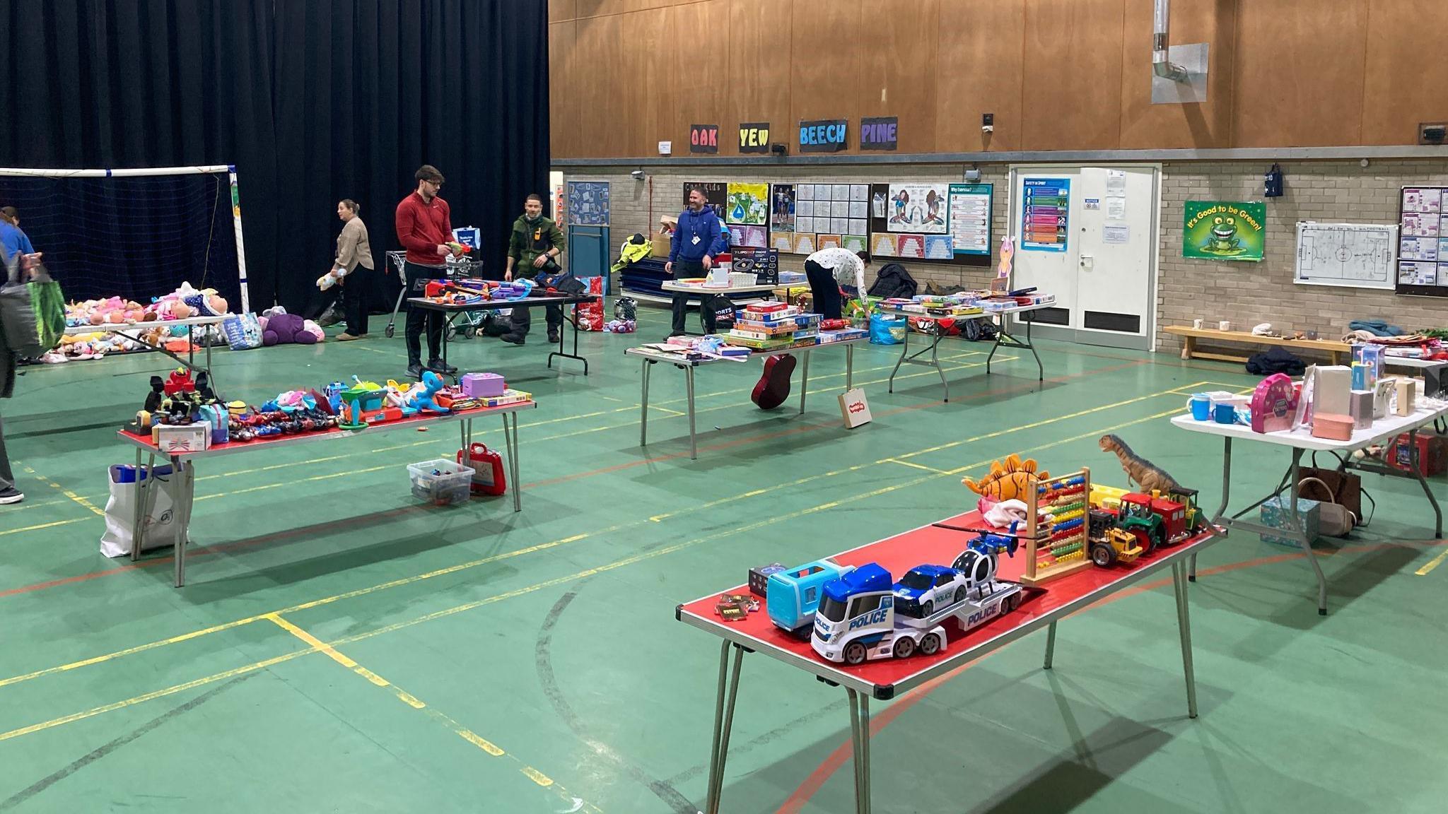 Tables of toys and gifts are set up in rows in a school sports hall. There are toy dinosaurs, trucks and cuddly toys. People are walking between the tables, setting them up.