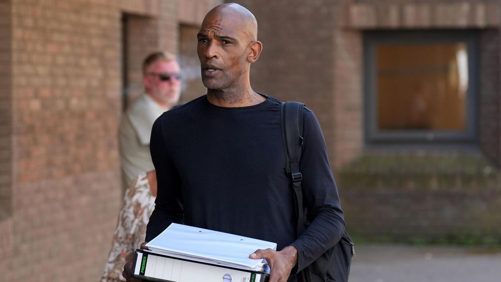 Matthew Martin wearing a black top and carrying two folders 